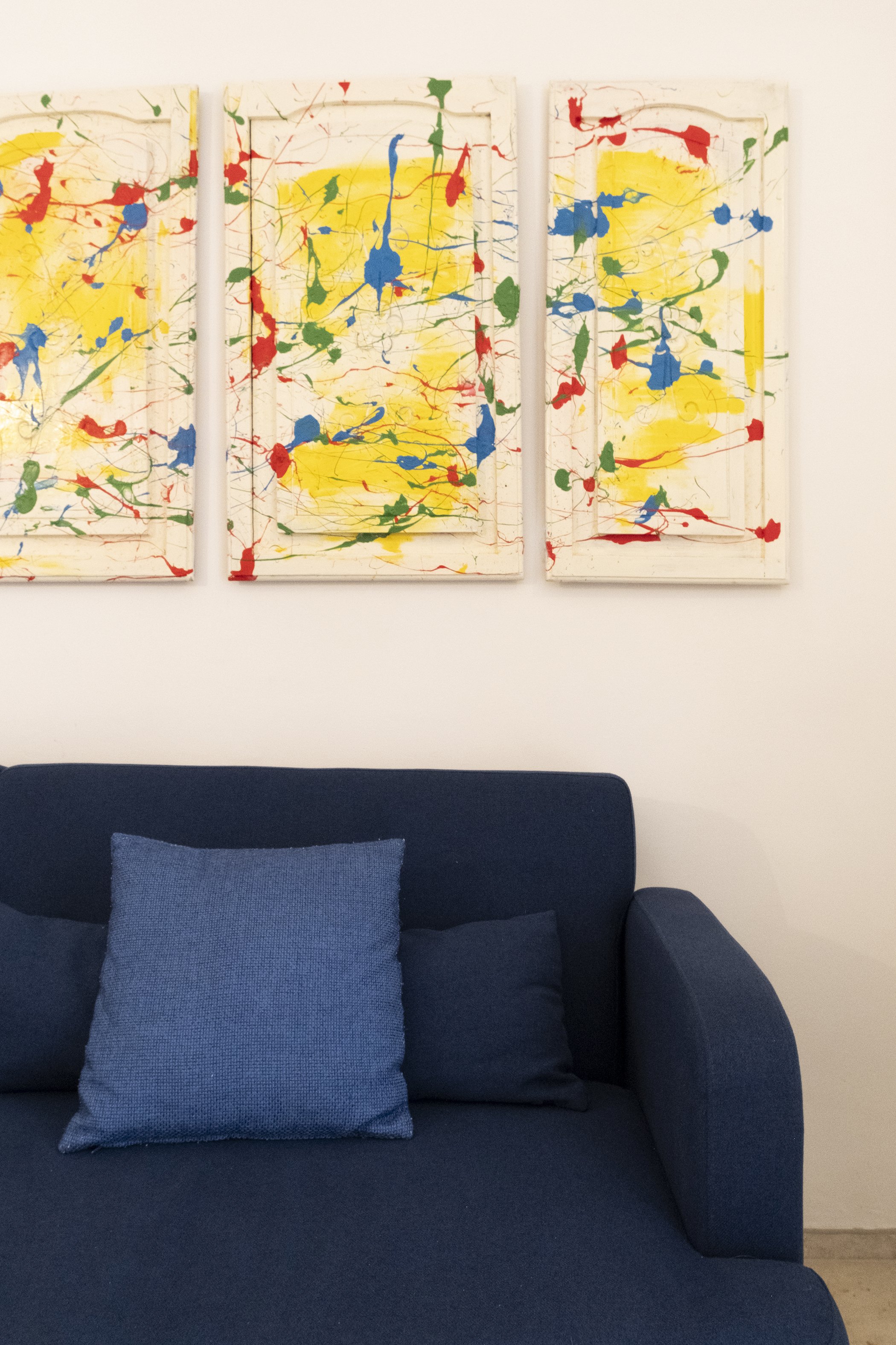 A blue sofa with dual blue throw pillows in front of an abstract triptych painting with splashes of red, yellow, green, and blue on white backgrounds.