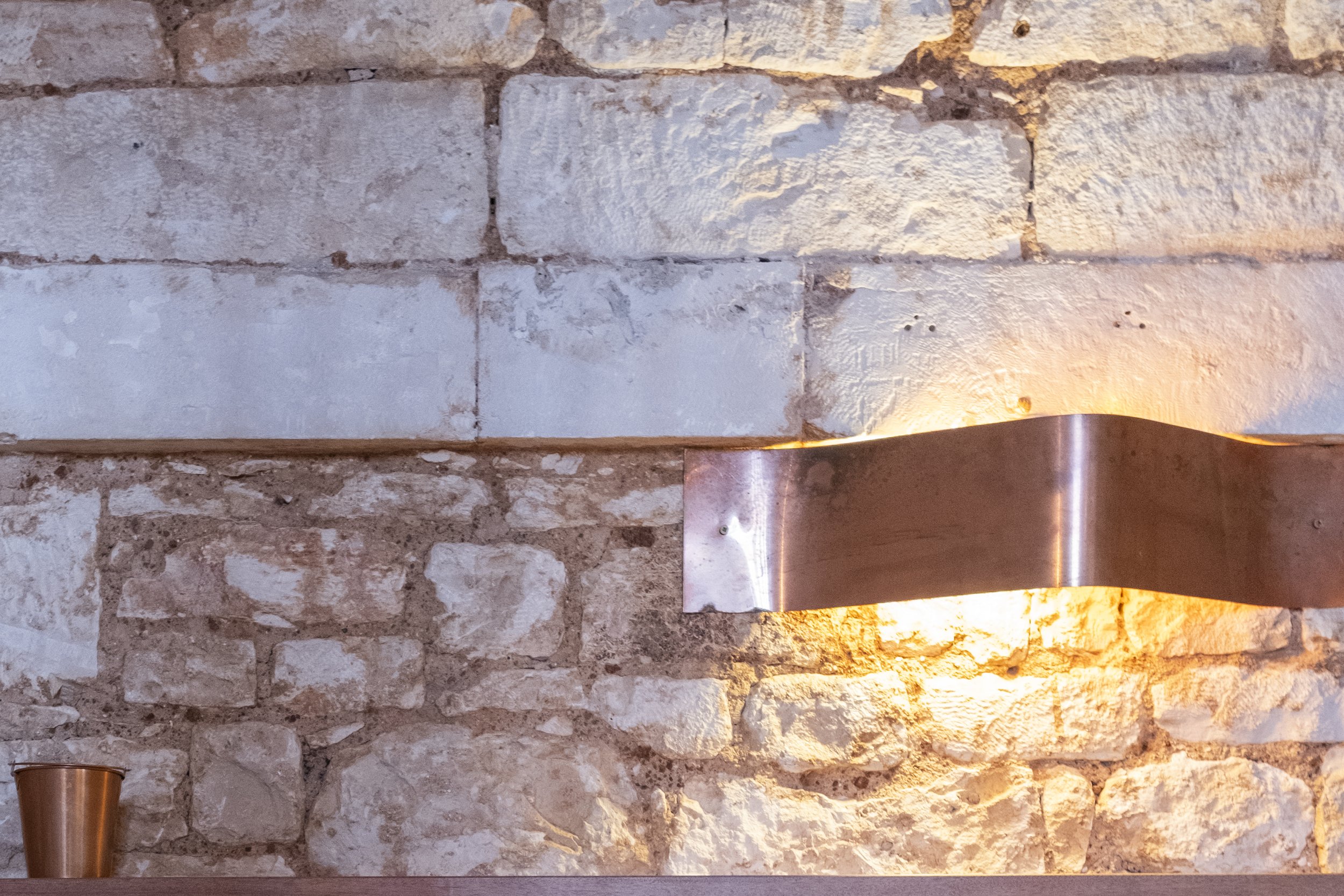 A stone wall with a modern, curved metal light fixture attached to it. The light casts a warm glow on the wall, and a copper-colored cup is placed on the surface below the light.