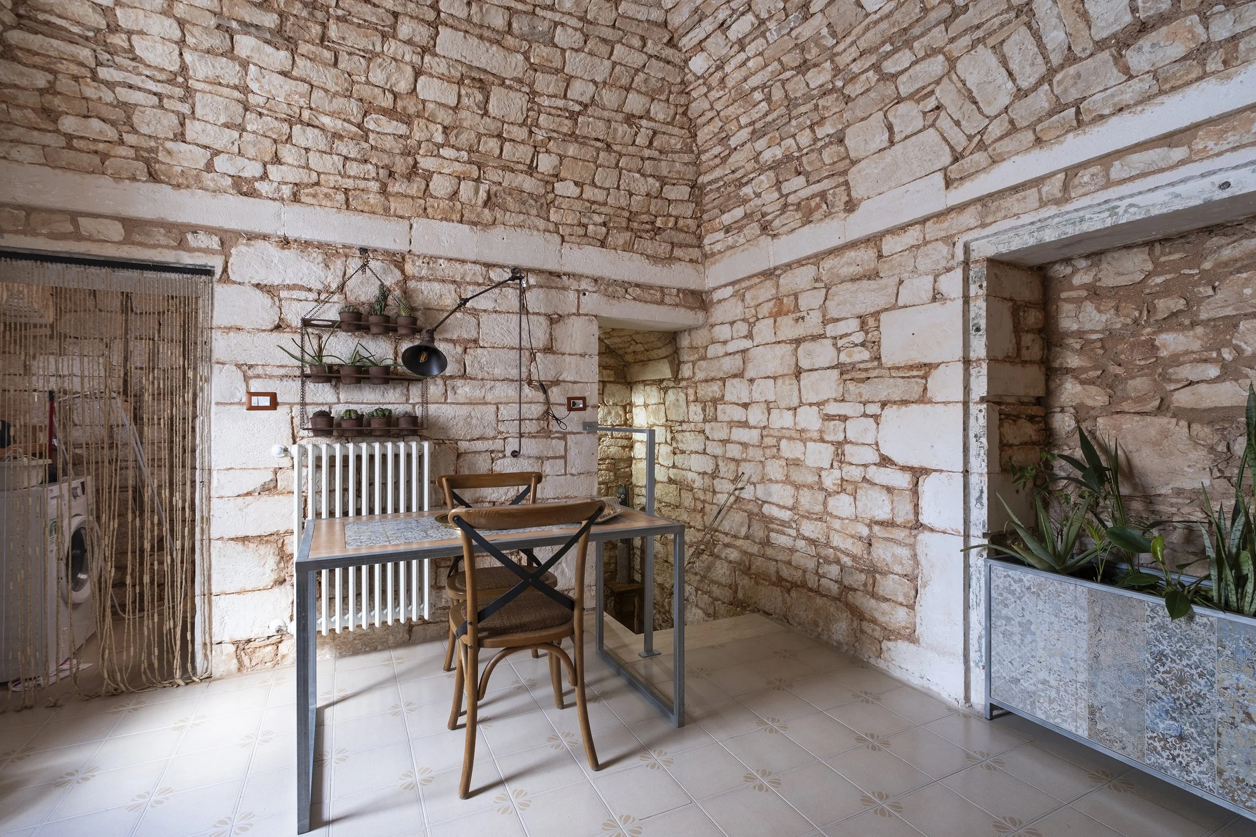 Room with exposed brick walls, small table with chairs, potted plants, and a decorative shelf.