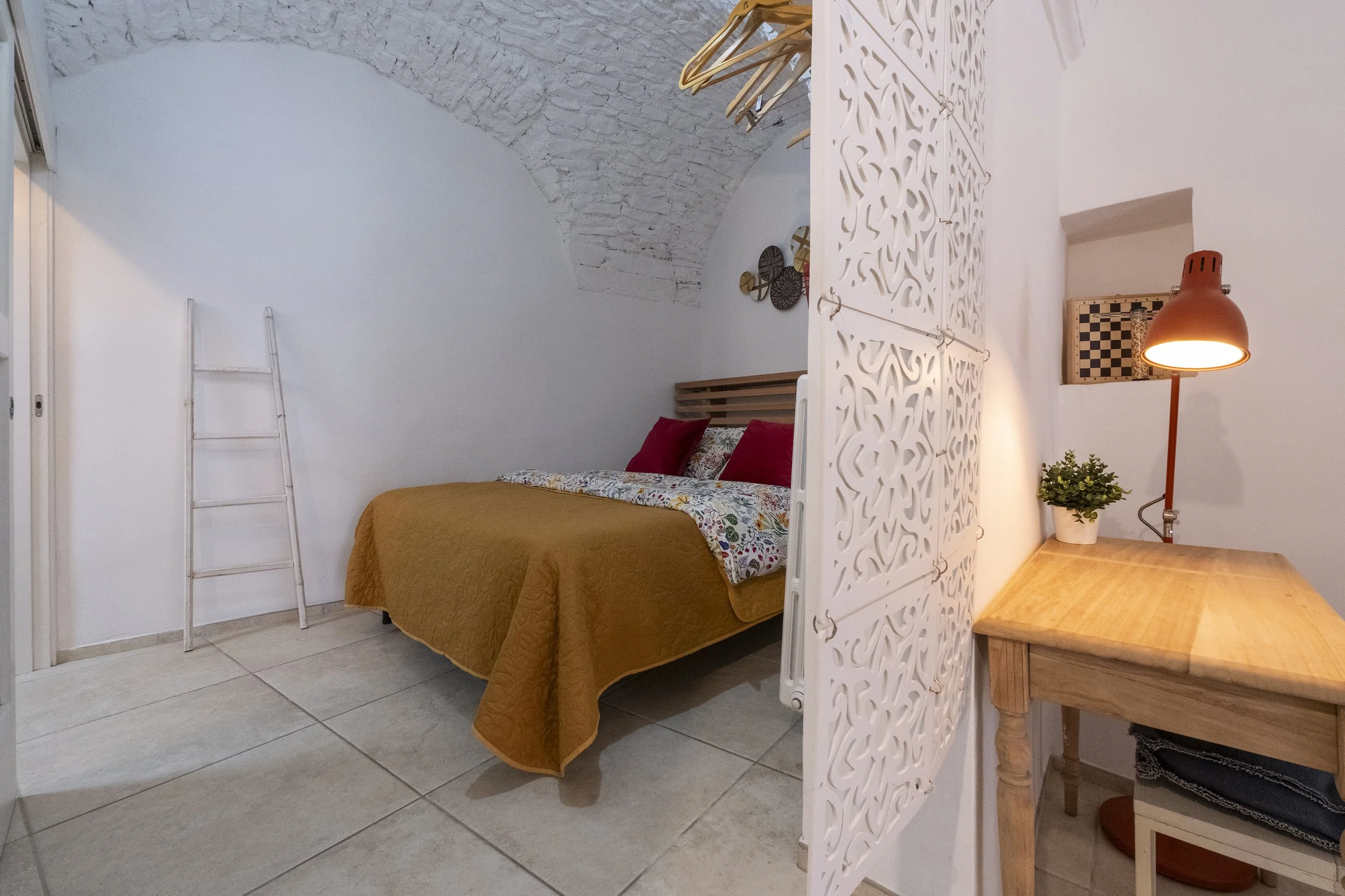 A cozy bedroom with a bed covered in a yellow quilt, colorful pillows, a white decorative room divider, and a wooden desk with a small green plant and a orange desk lamp.