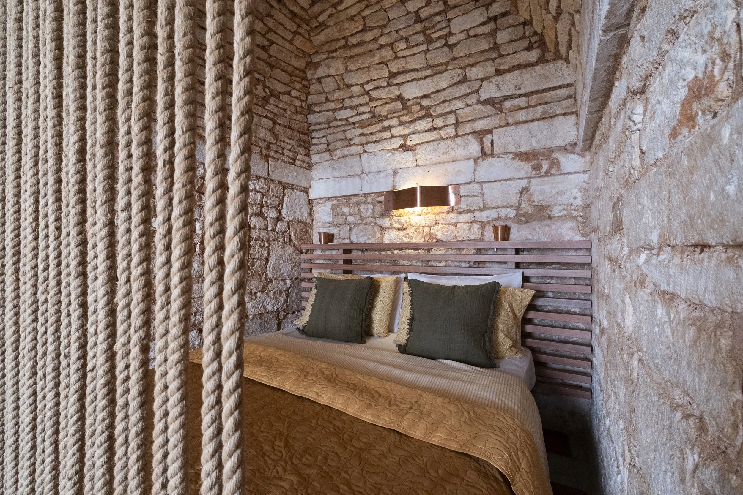 A bedroom with stone brick walls and a wooden bed frame with green and patterned pillows, a wall-mounted copper light fixture, and decorative copper cups on the headboard.