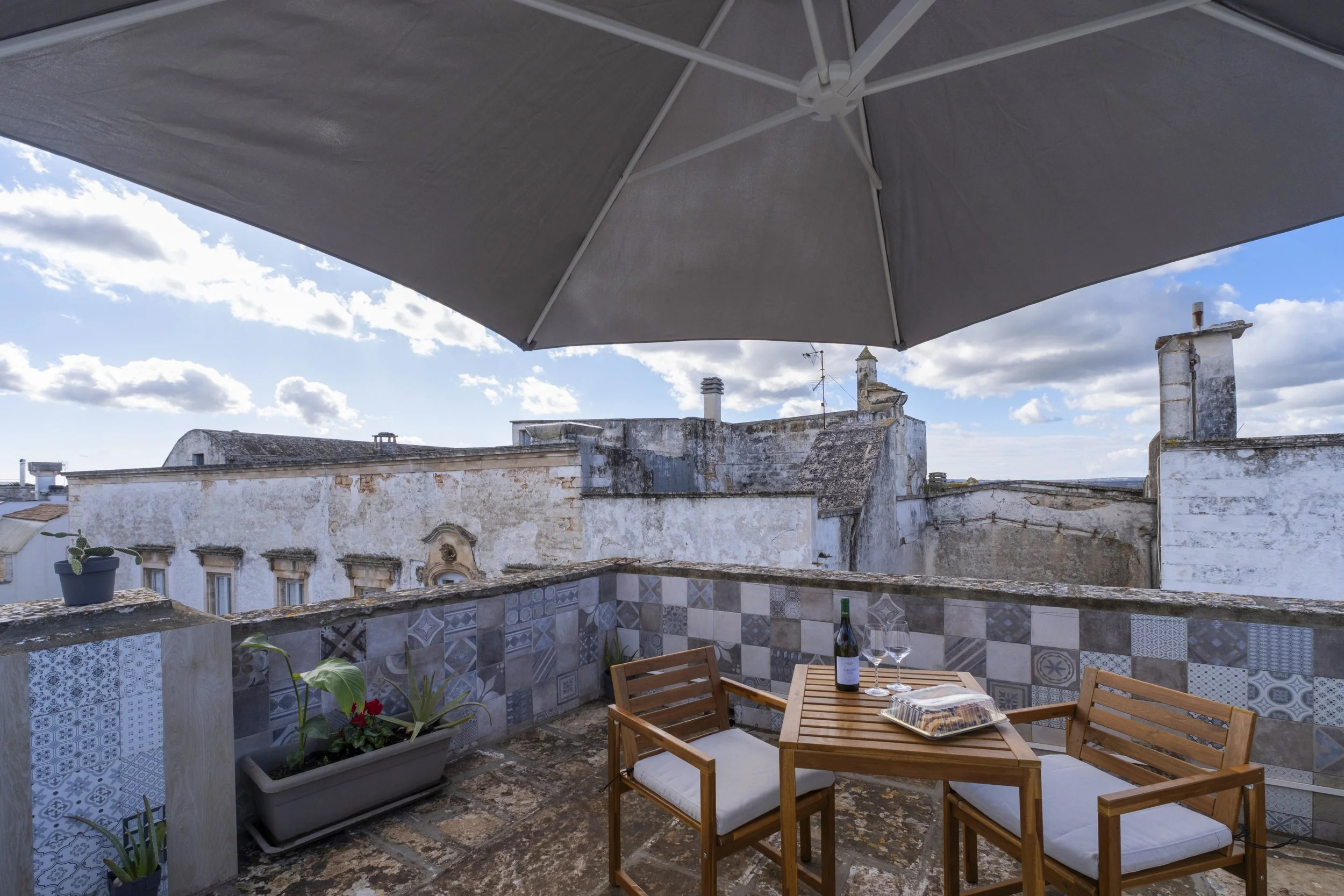 An outdoor rooftop patio with a wooden table, two chairs with white cushions, wine bottle, glasses, and a pizza box, under a large grey umbrella, overlooking weathered white buildings and a partly cloudy sky.