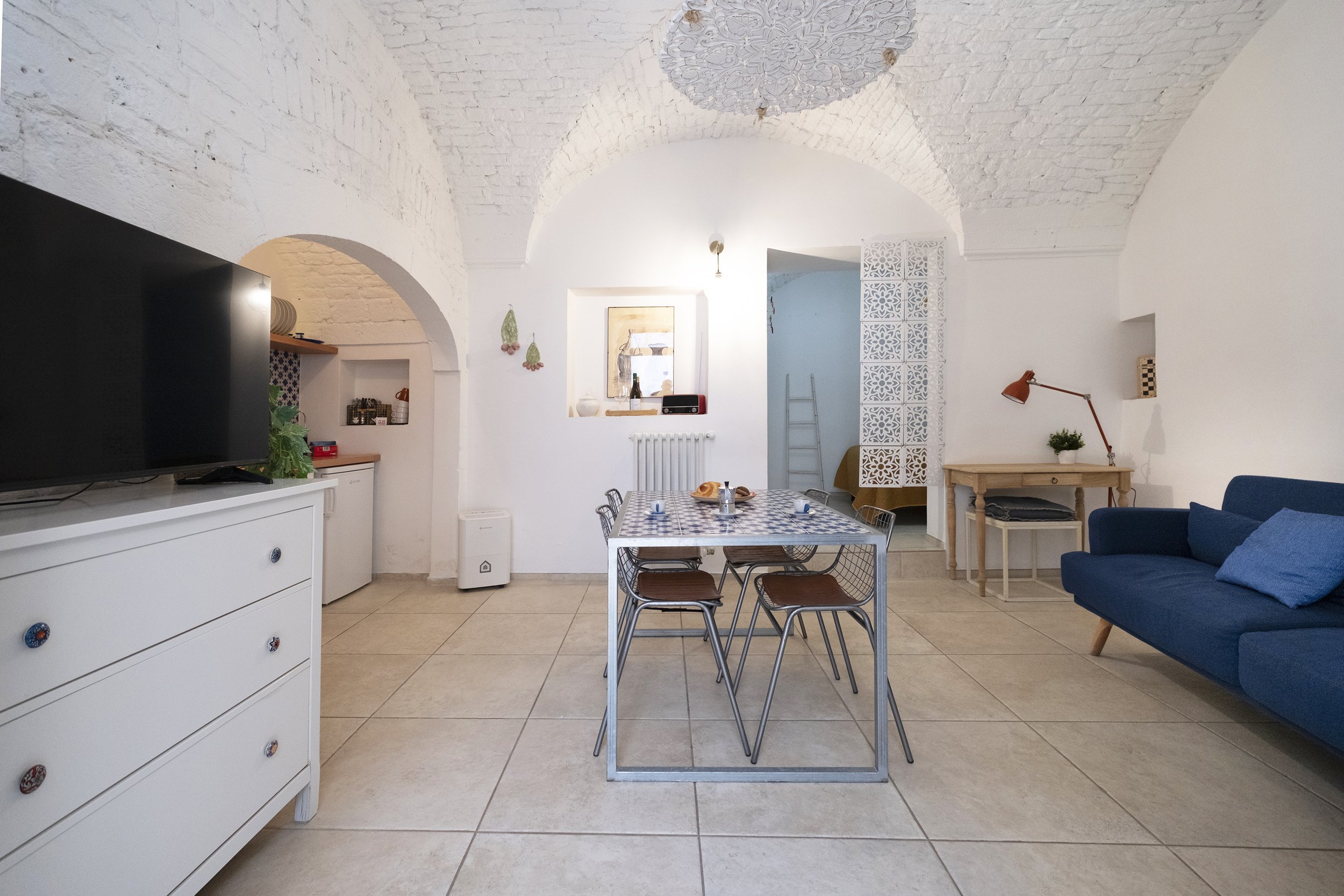 A bright living room with a dining table, blue sofa, and vintage decor. There are arched white brick ceilings, a white dresser with a TV, and a small kitchen area in the background.