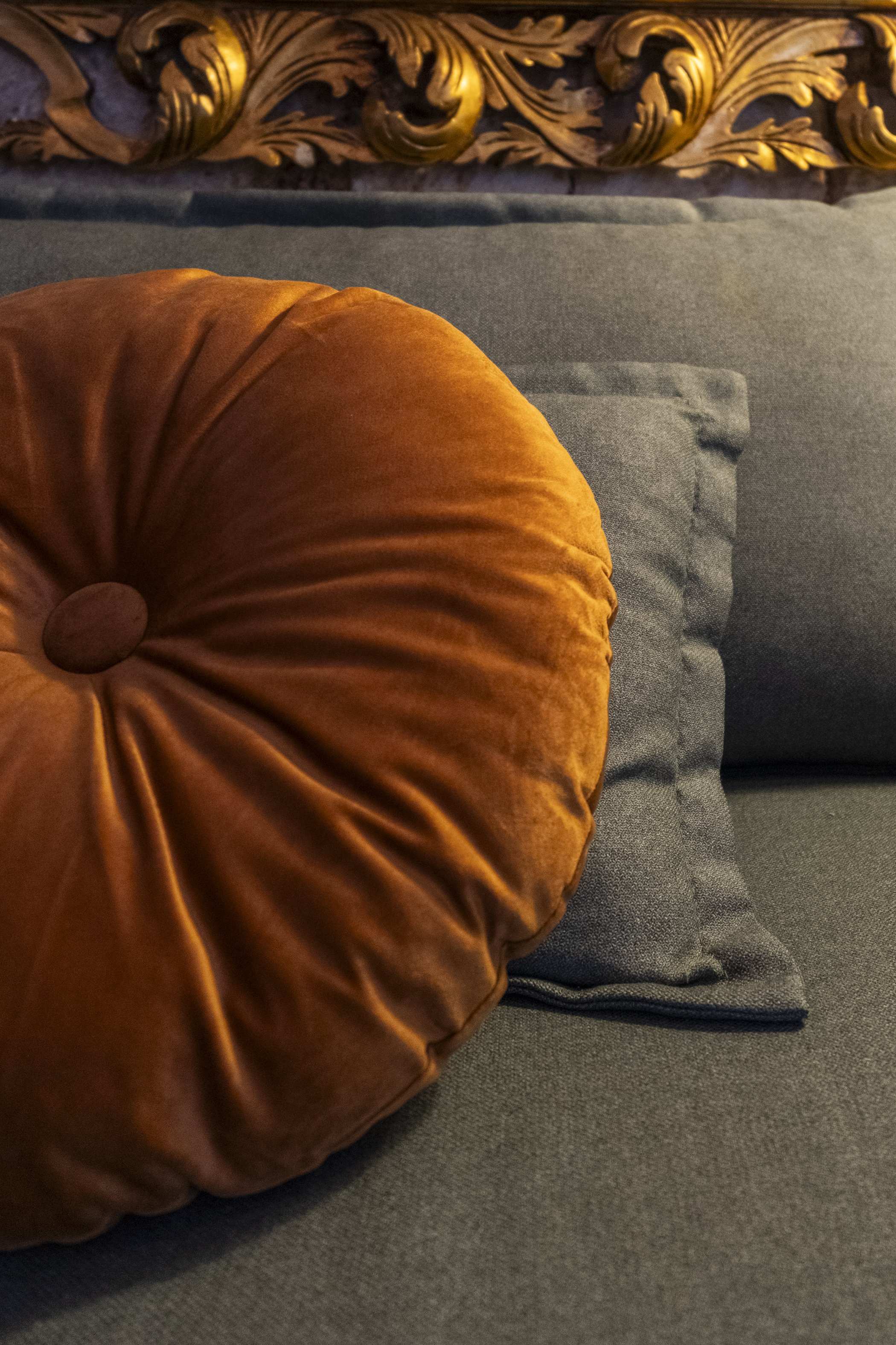 Close-up of a round, orange velvet pillow on a gray fabric sofa, with ornate gold carved wooden decoration in the background.