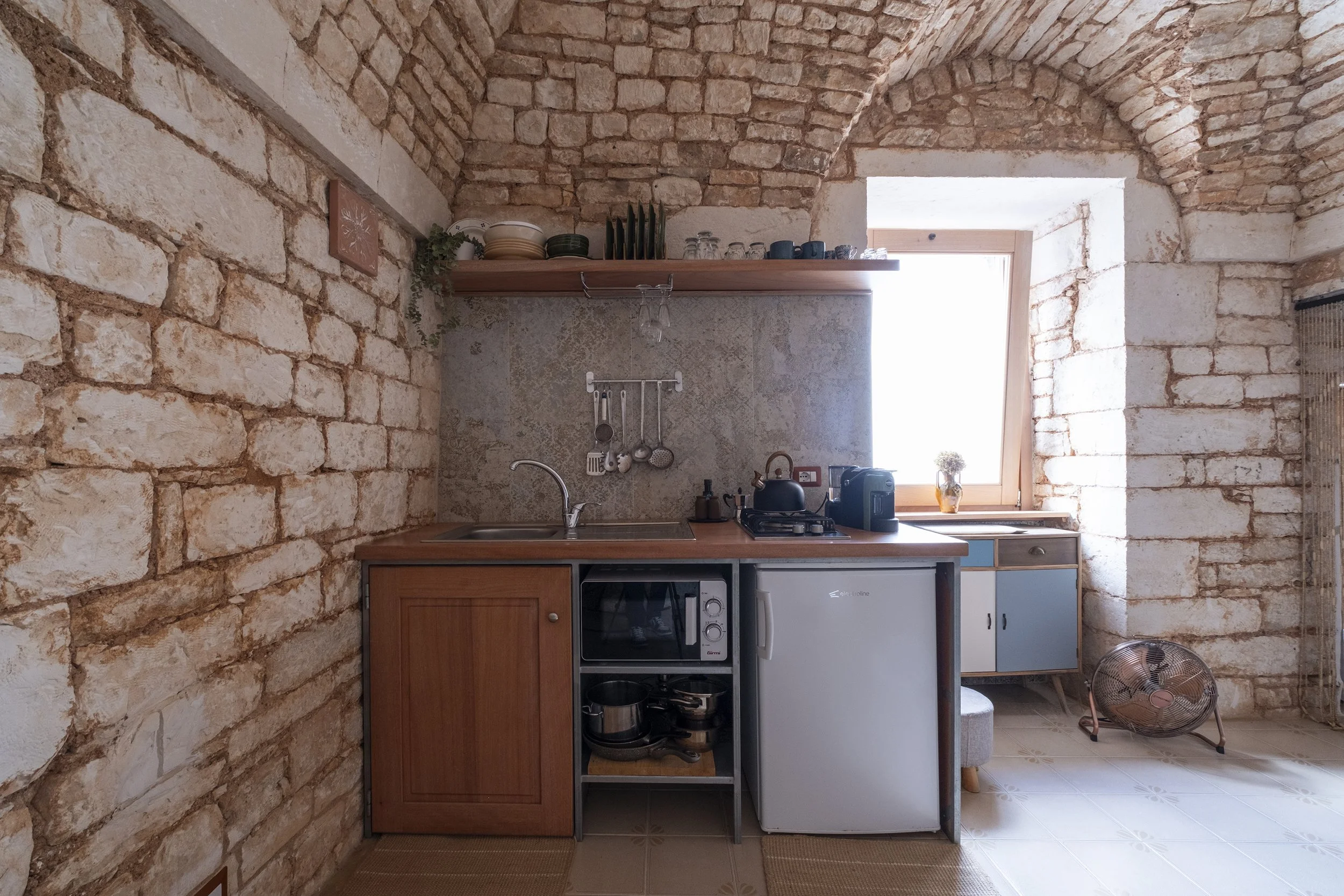 A rustic kitchen with stone brick walls, a small sink, and a wooden countertop. Above, a wooden shelf holds dishes and glasses. A window lets in natural light, and a small counter area has a teapot, toaster, and small fridge. There is a fan on the fl