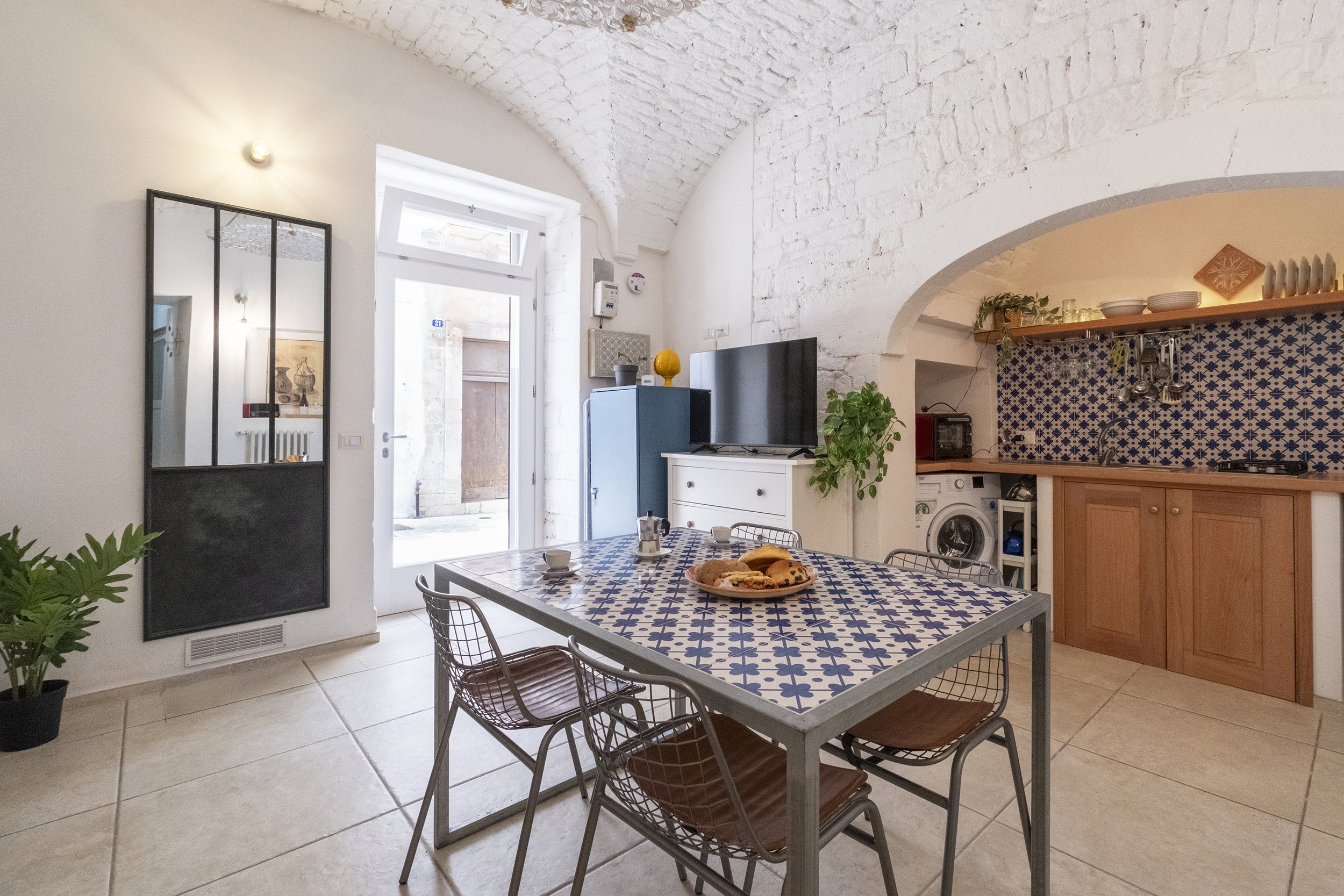 A dining area with a table covered in blue and white patterned tiles, surrounded by metal chairs with brown seats. There is a plate of baked goods on the table, and the room features white brick arches and walls, with an open kitchen area in the back
