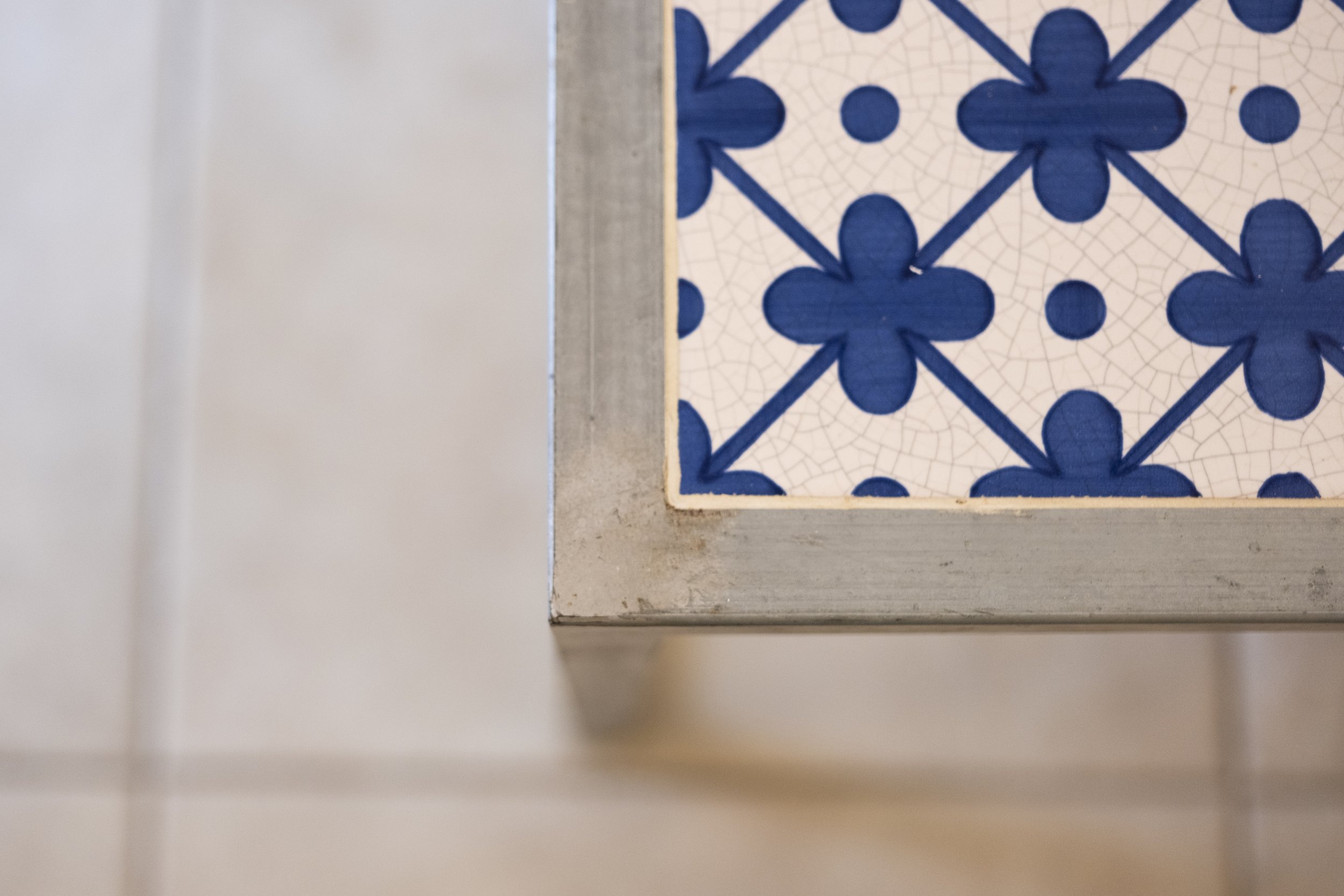 Close-up view of a decorative blue and white tile on the corner of a white painted surface, with visible grout lines and a rustic wooden border.