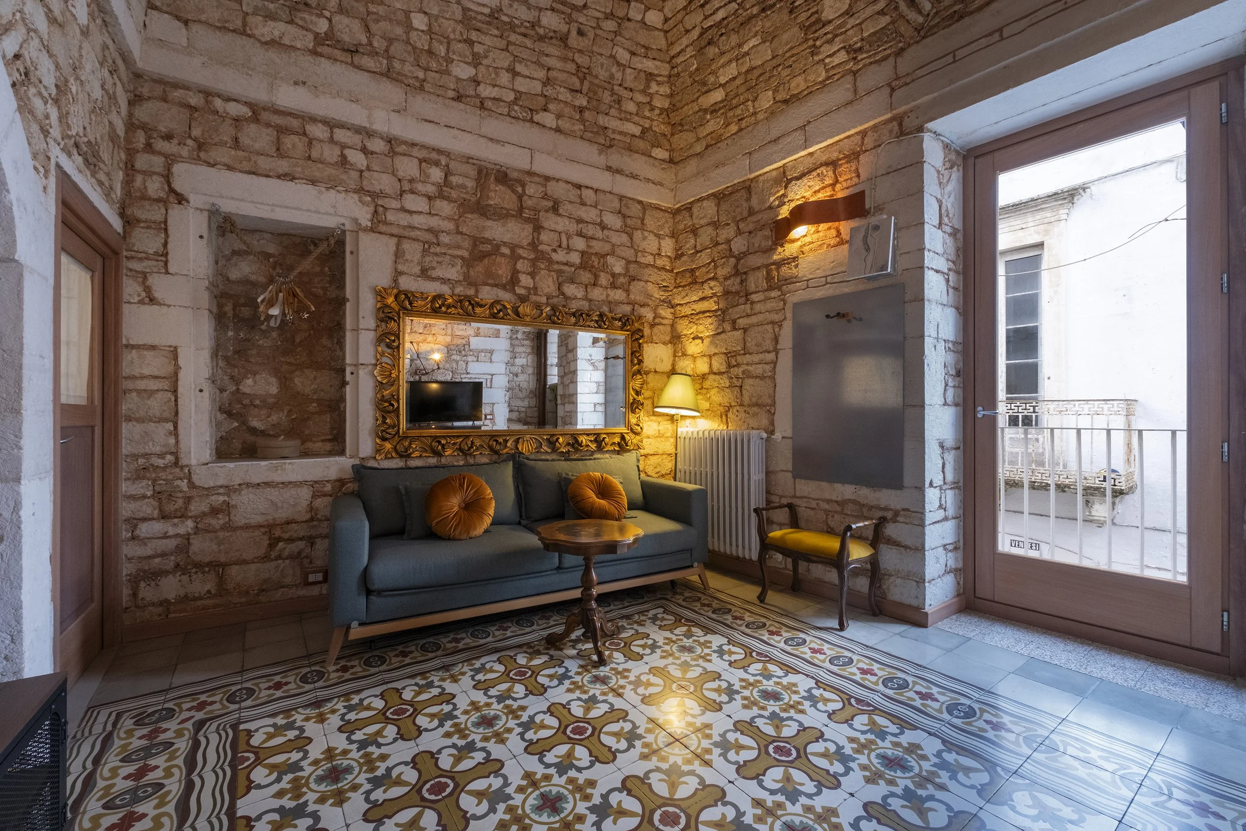 Living room with exposed brick walls, a vintage mirror, a teal sofa with orange pillows, a small wooden coffee table, a chandelier reflection in the mirror, a standing lamp, a small wooden armchair, and a glass door leading outside.