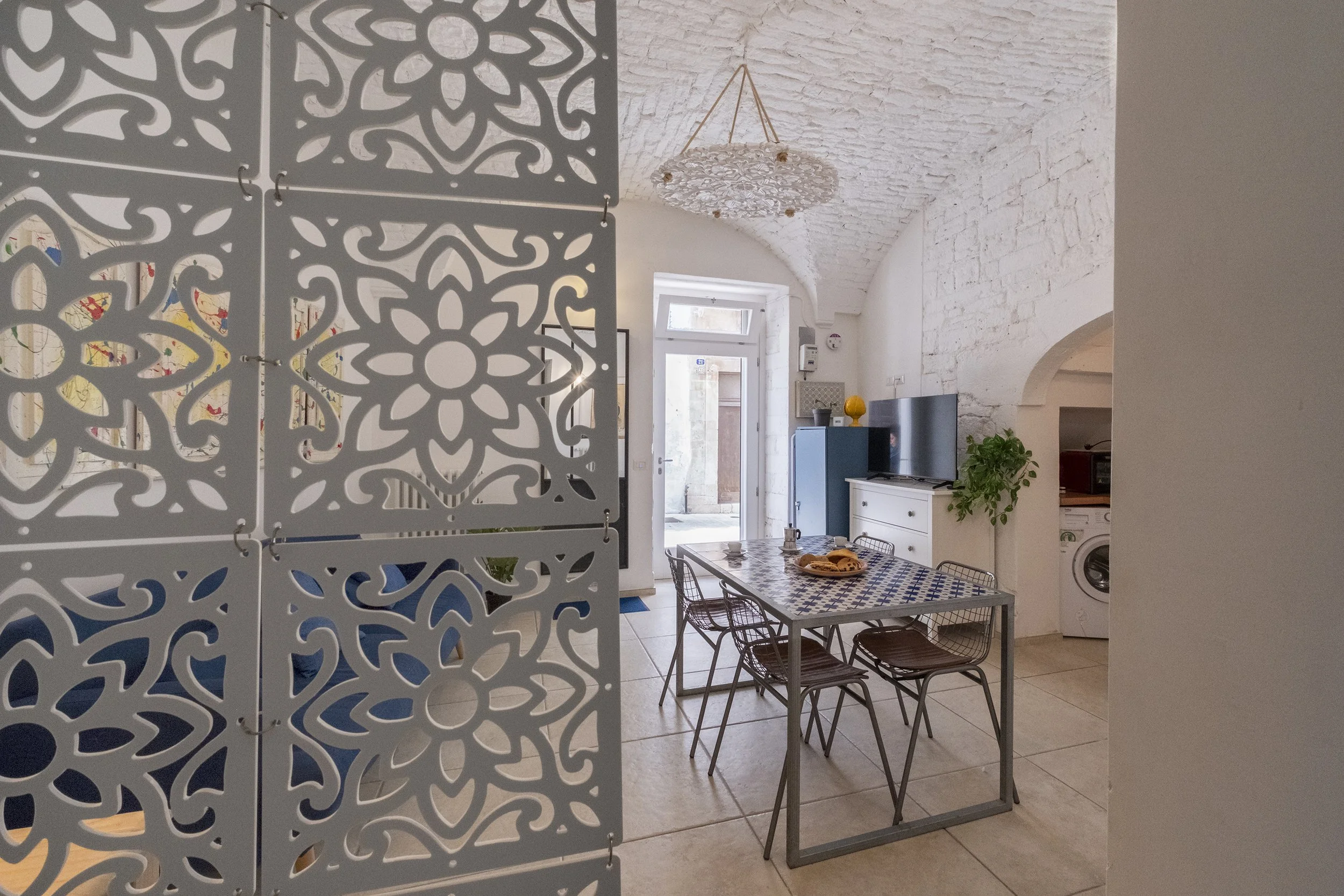 Interior of a bright living room with white brick walls, a dining table with a patterned tablecloth, chairs, a TV, a blue cabinet, a hanging light fixture, and a washing machine in a small nook.