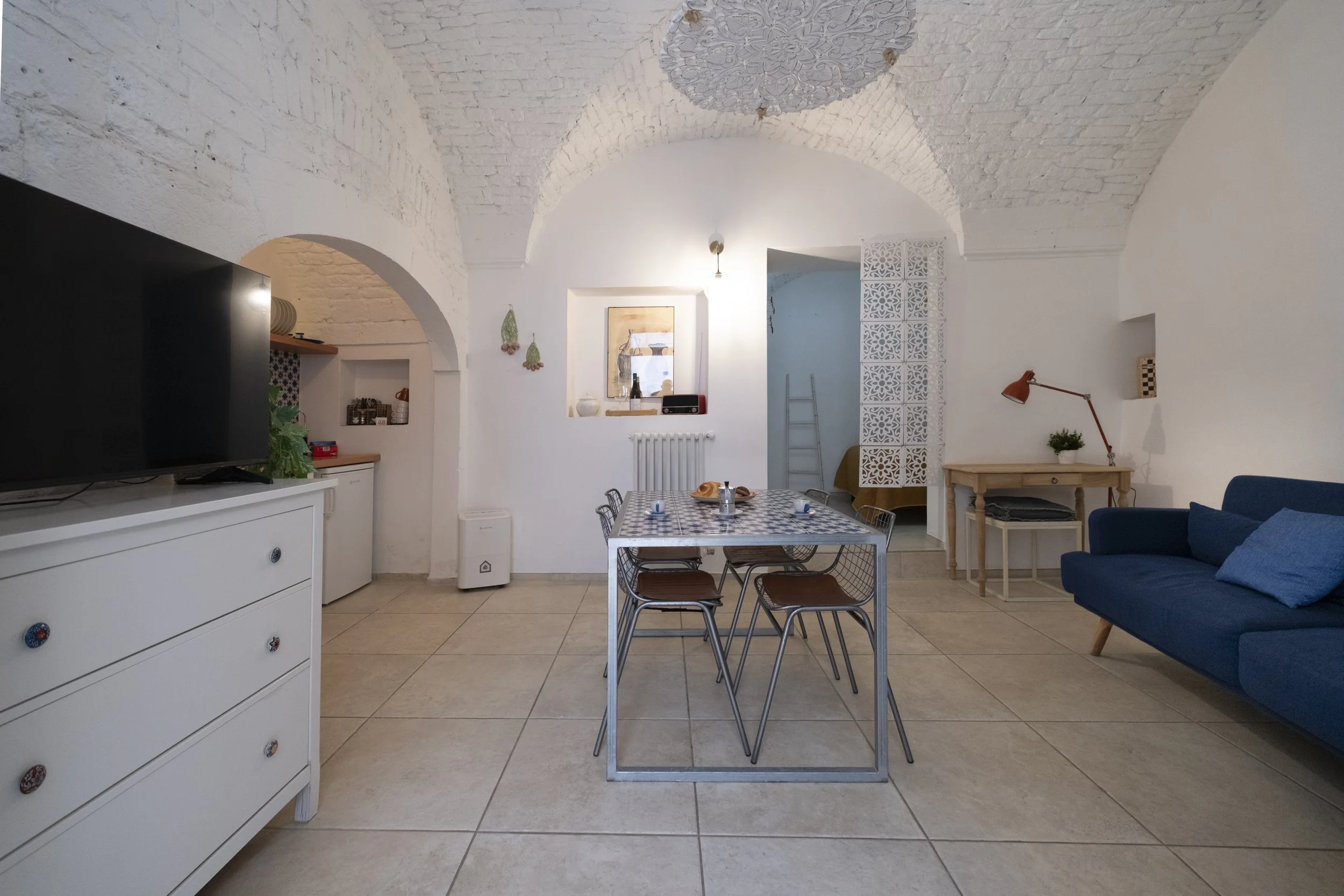 A living room with white painted brick arched ceilings, a blue sofa, a dining table with a patterned tablecloth and six chairs, a white dresser with a TV on top, and a small corner kitchenette. The room is decorated with small hanging baskets, a fram