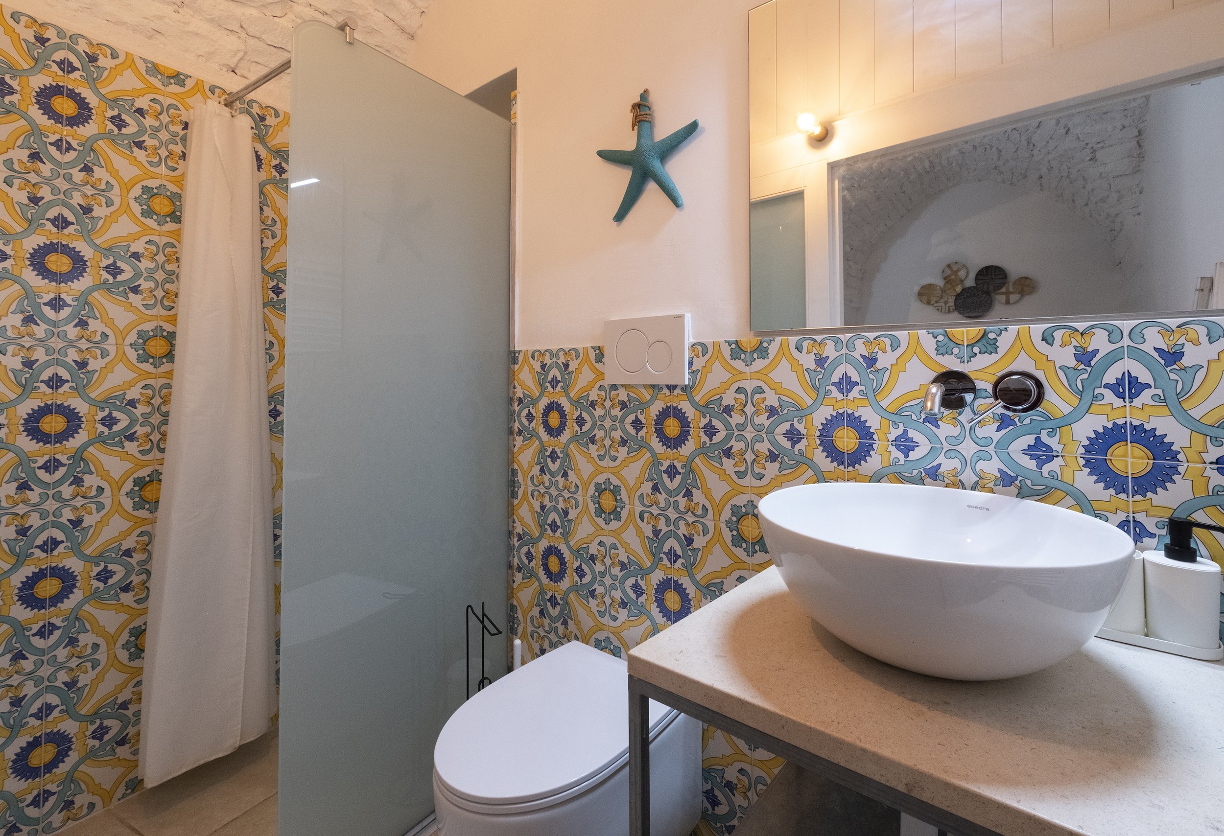 Bathroom with patterned yellow, blue, and green tiles on the wall, a starfish decoration on the wall, a white vessel sink on a beige countertop, toilet, shower with frosted glass door, and a mirror.