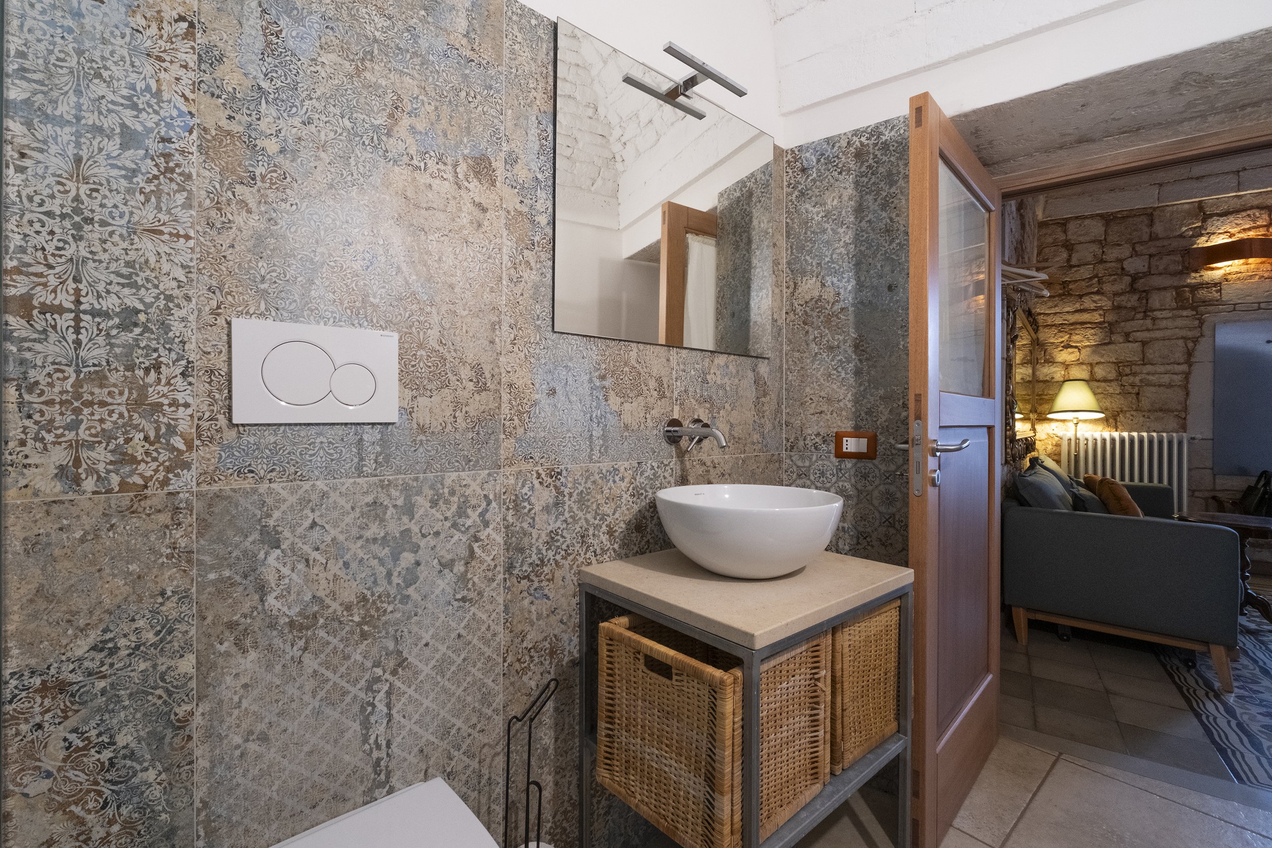 Small bathroom with textured, patterned stone tiles, a round white vessel sink on a countertop, a large mirror above, and a partially open wooden door leading to a living room with brick walls, a sofa, a floor lamp, and a radiator.