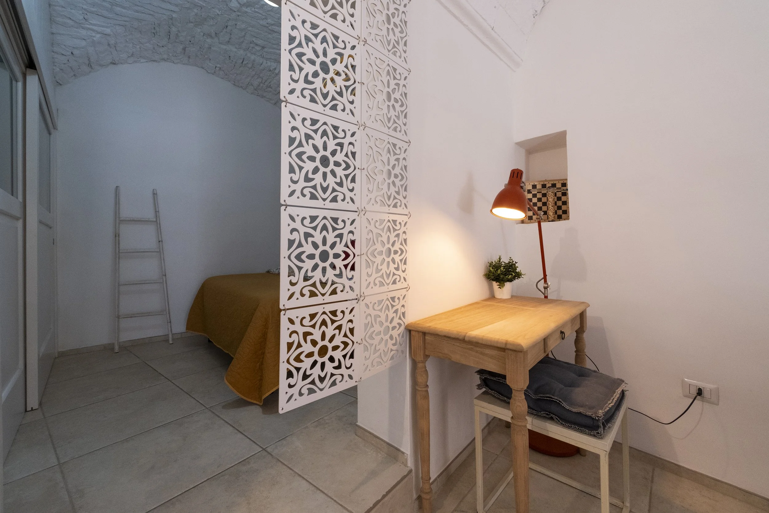 Corner of a room with a small desk, a red desk lamp, a potted plant, and a pile of folded clothes, with a bed in the background separated by a decorative white partition and a ladder leaning against the wall.