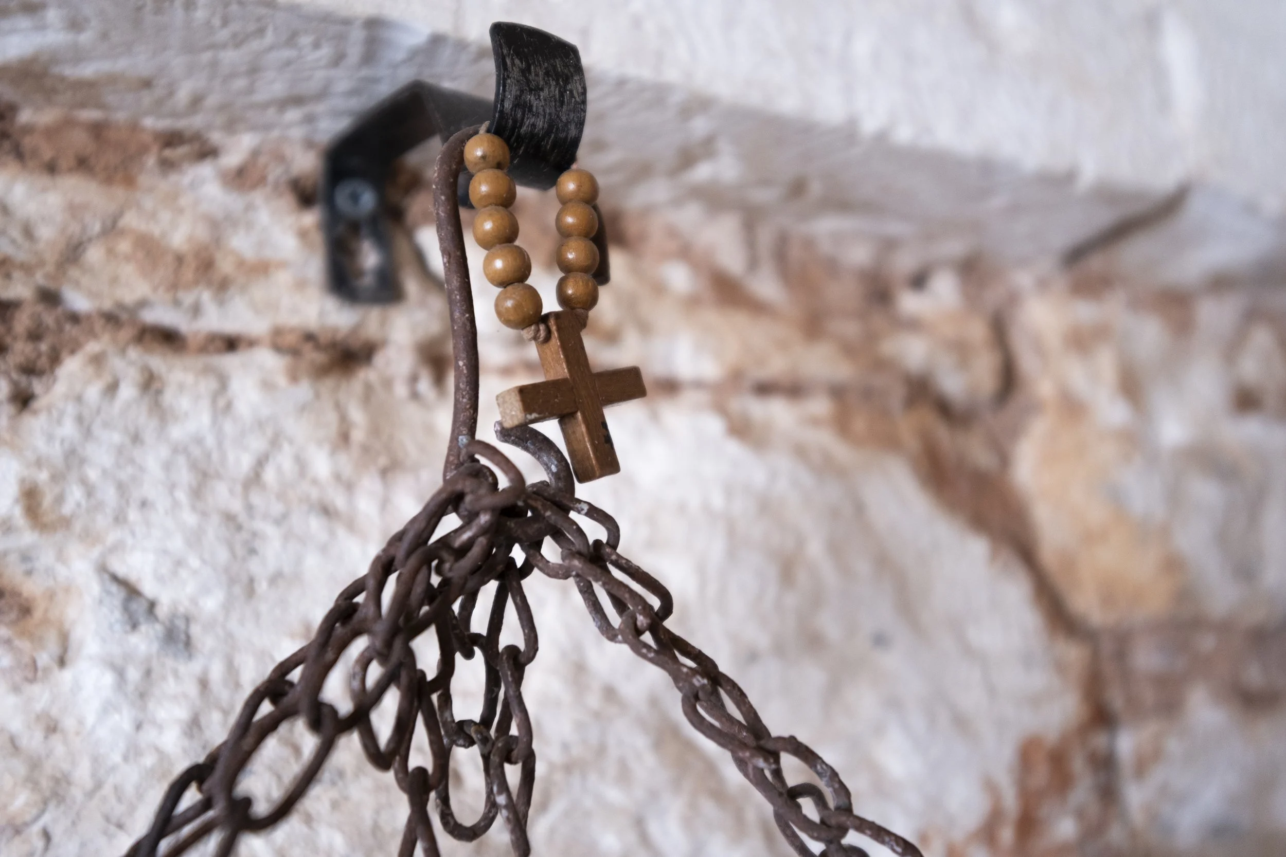 A rusty chain hanging from a black hook on a stone wall, with a wooden rosary with wooden beads and a cross resting on the chain.