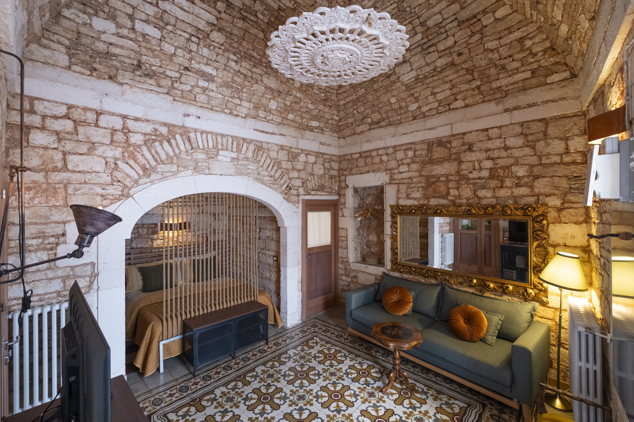 A cozy living room with exposed brick walls, a green sofa with two round brown pillows, a large ornate gold mirror, a small wooden coffee table, a vintage-style floor lamp, and a patterned tile floor. There is a bed partially visible through an arche