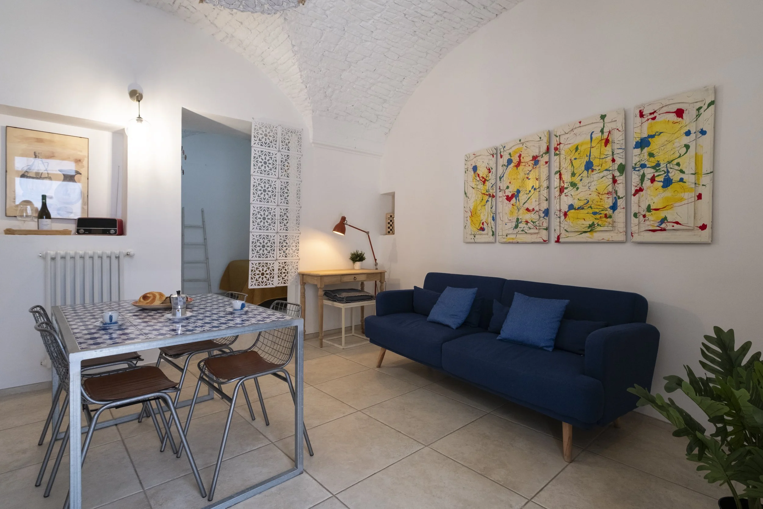 Living room with a blue sofa, colorful abstract paintings on the wall, a wooden table with a lamp, and a dining table with six chairs and breakfast items.