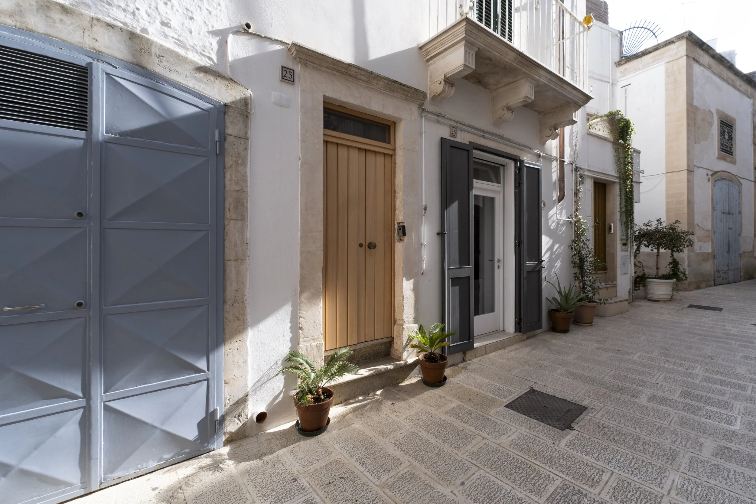 Residential street with white buildings, multiple doors, potted plants, and a large gray metal gate.
