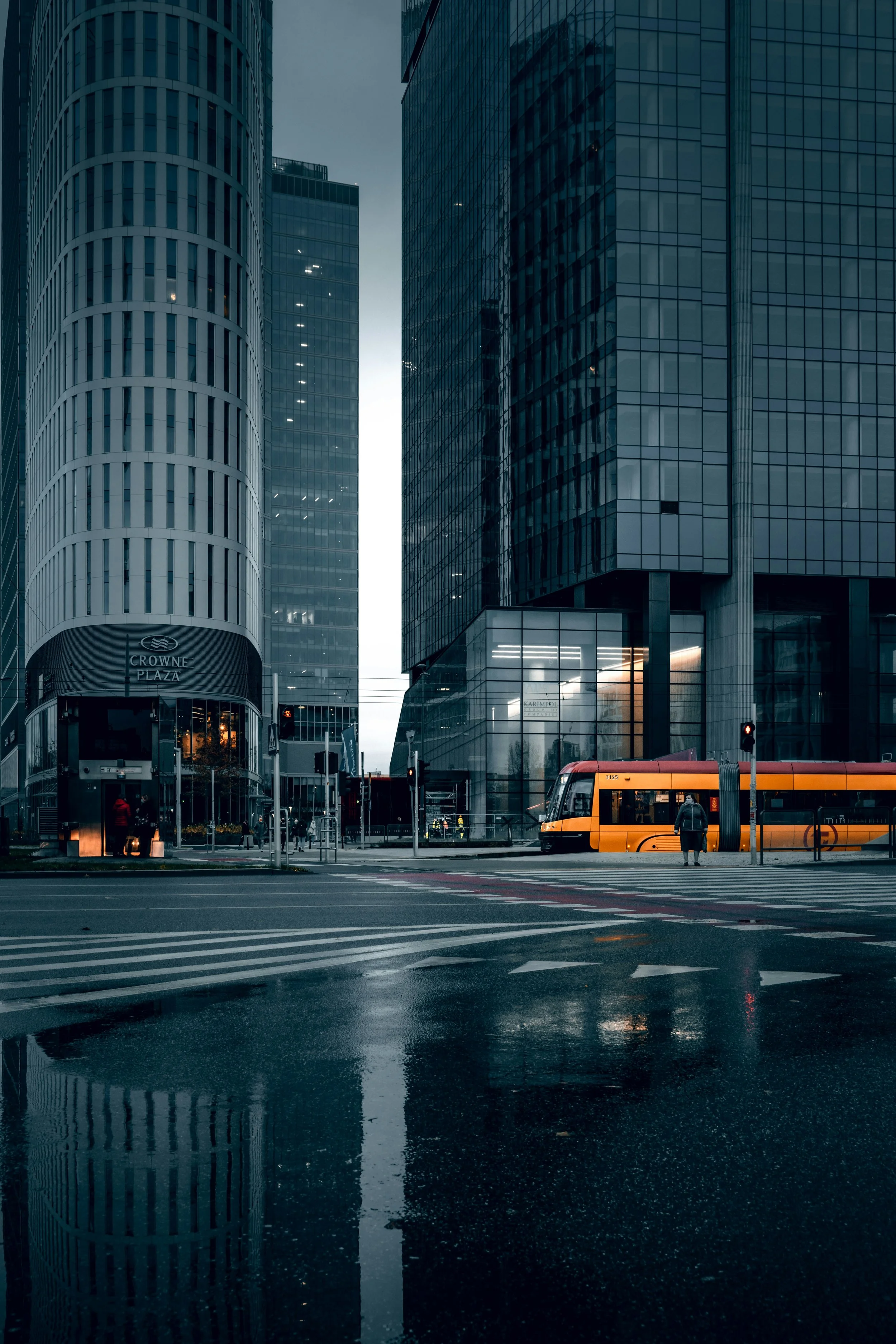 A city street with modern glass skyscrapers, a yellow-orange tram, and a solitary pedestrian on a rainy day.