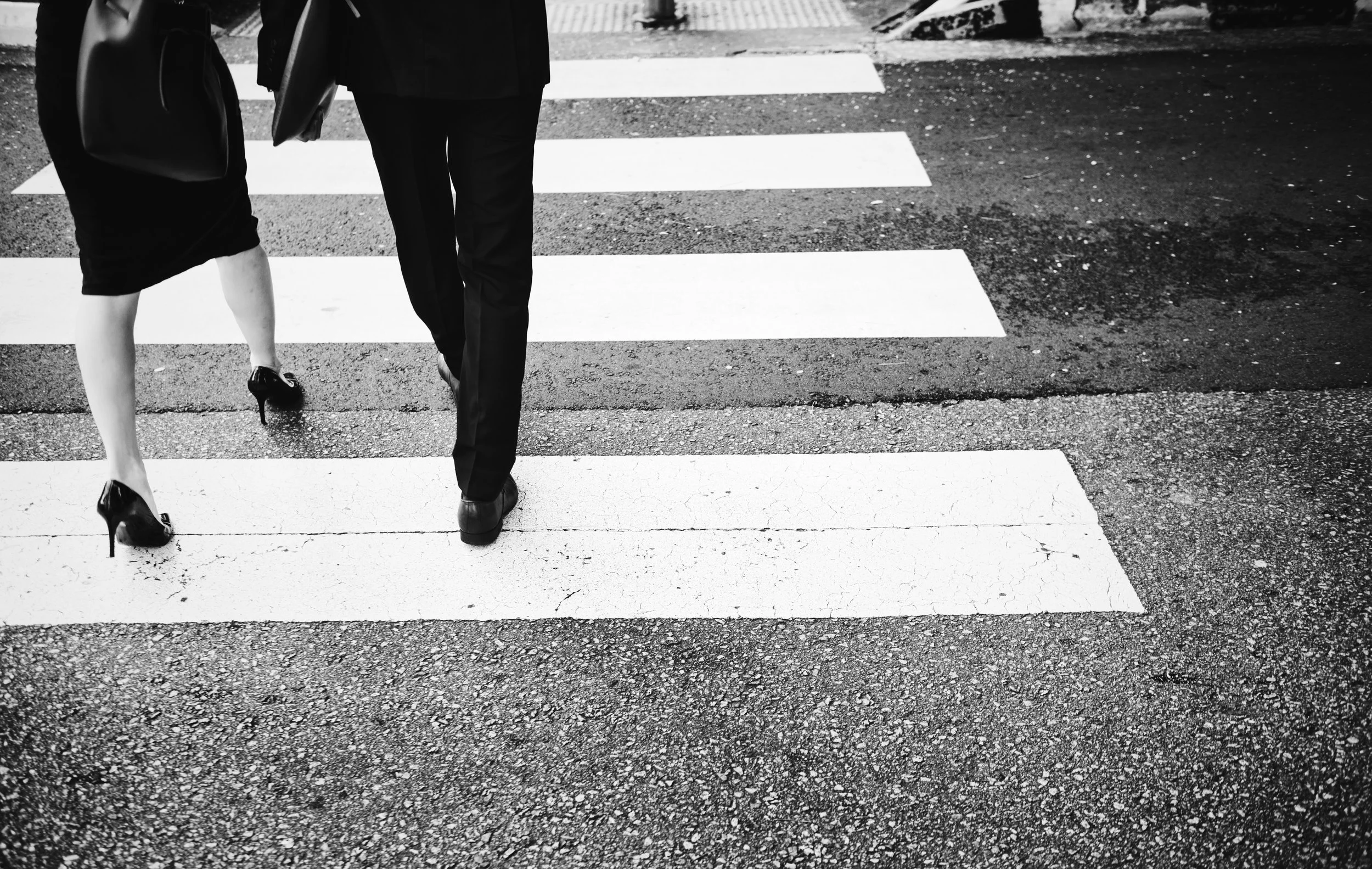 Two people walking across a pedestrian crossing, one in a skirt and heels, the other in pants and shoes, black and white photo.
