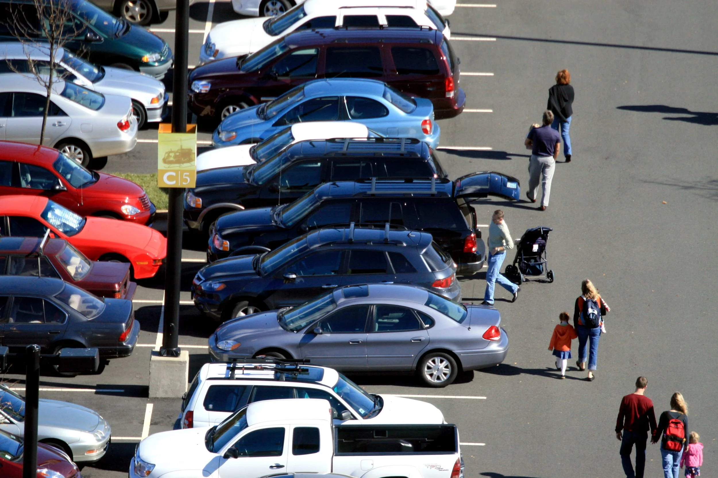 Top-down view of a parking lot with various parked cars and people walking, including a woman pushing a stroller and children with adults.