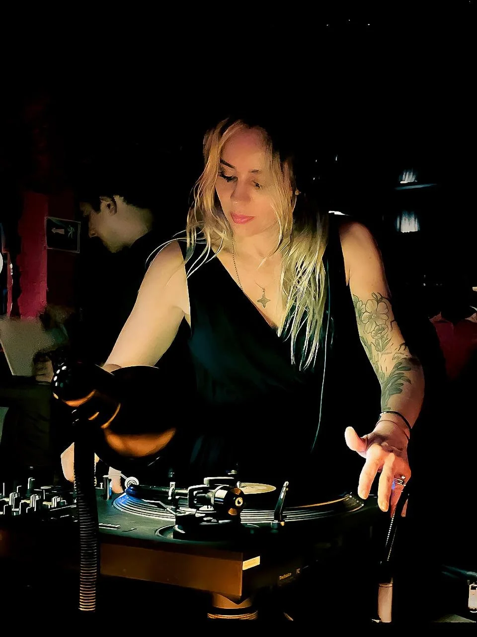 Mujer con cabello rubio y tatuajes en el brazo operando una tornamesa en un ambiente oscuro.