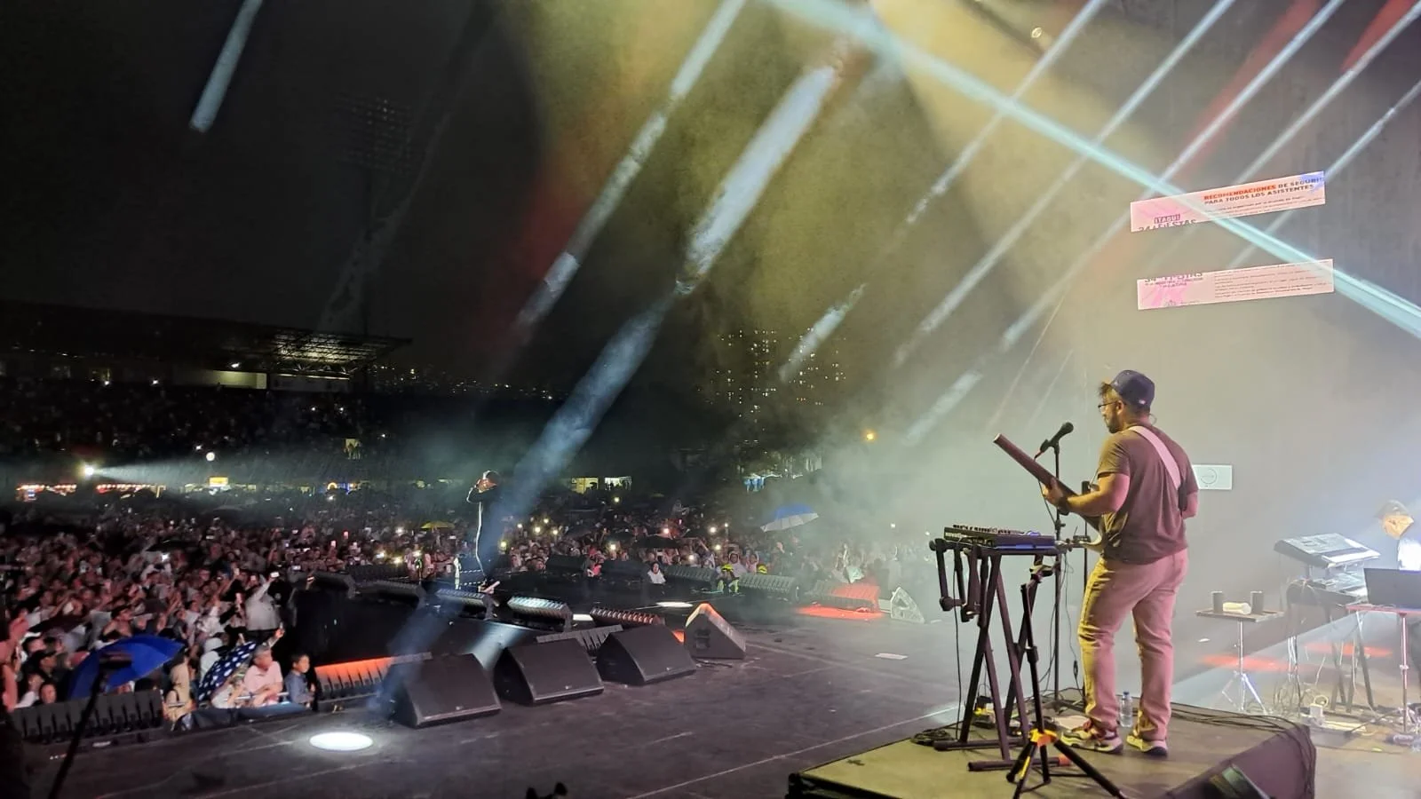 Un concierto al aire libre con un artista tocando la guitarra eléctrico en un escenario, frente a una multitud de personas bajo la noche, con luces de escenario y un ambiente festivo.
