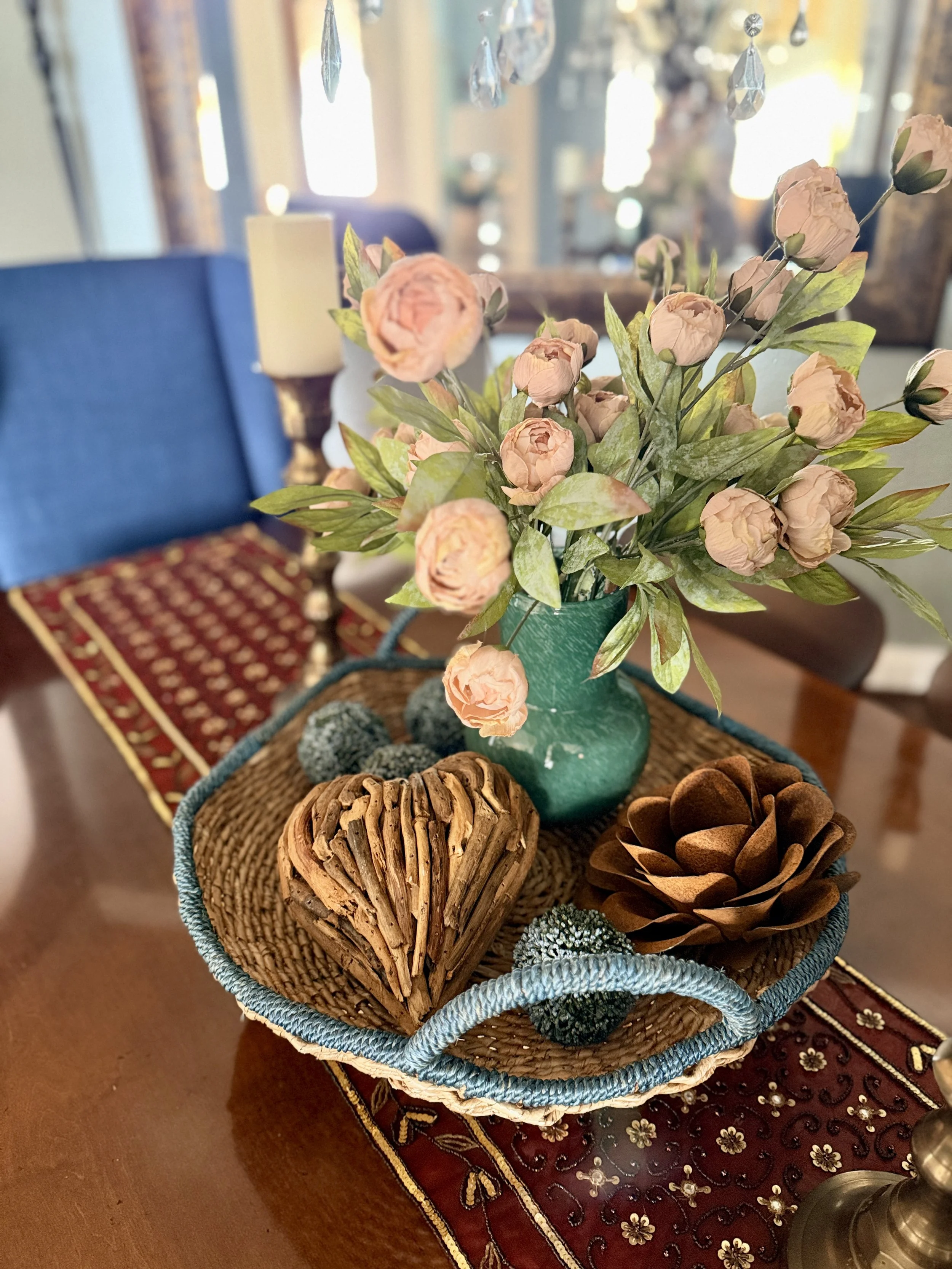 Styled vignette with blush flowers in a green vase on a woven tray, layered with other collected items in a warm curated home.