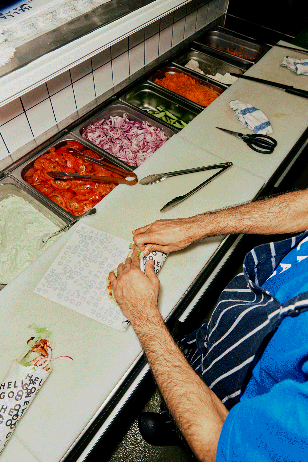 A worker wraps a pizza slice in branded paper at a pizza restaurant counter with various fresh vegetables in metal containers, including tomatoes, onions, cucumbers, carrots, and vegetables in the background.