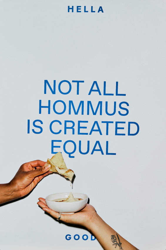Two hands holding a bowl and a piece of bread, with a sign in the background that reads 'Hello, Not all hummus is created equal. Good.'