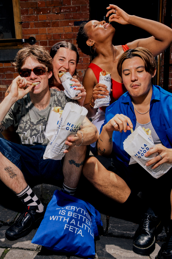 Four young people enjoying souvlaki wraps and drinks outside against a brick wall at the Hella Good Greek food restaurant, with one woman drinking from her hand and three smiling or making faces.