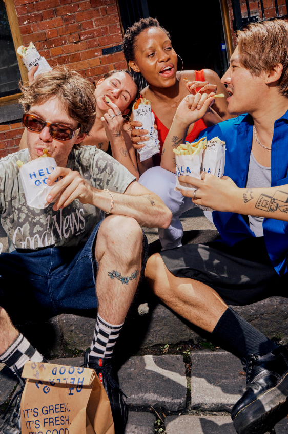 Four diverse young adults enjoying souvlakis and fries outside a brick building which is the Hella Good Greek food restaurant, sitting on a sidewalk.