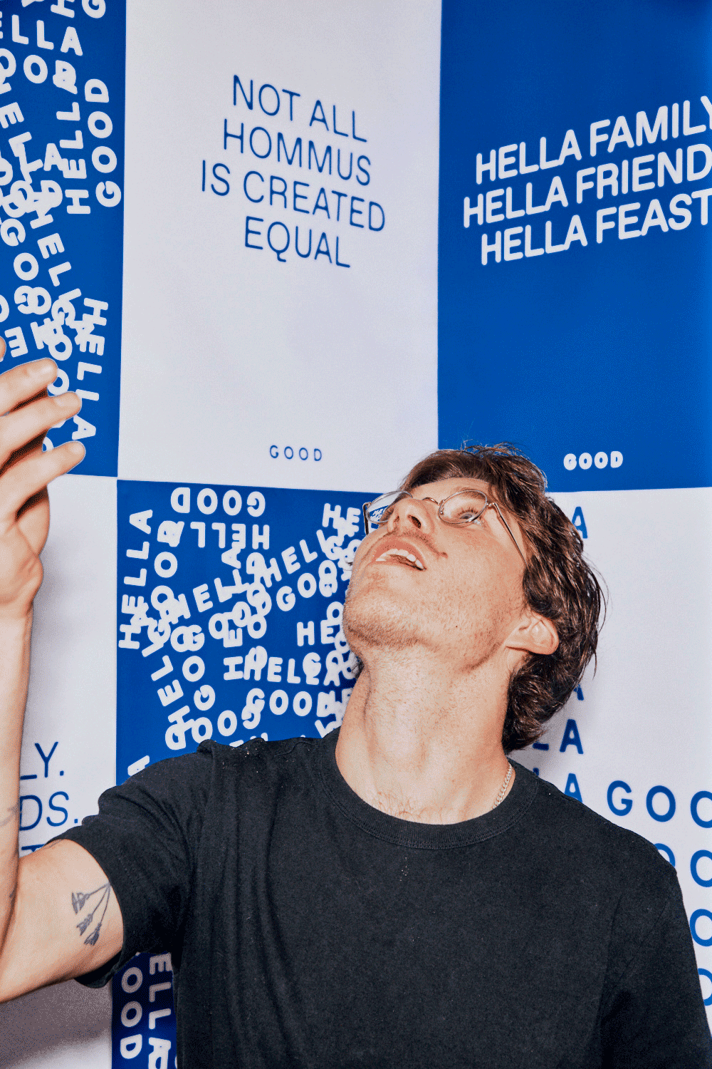 A man with glasses eating chips in front of a wall decorated with blue and white posters that have witty phrases about humans and families, including "Not all hommus is created equal" and "Hella family, hella friends, hella feast."