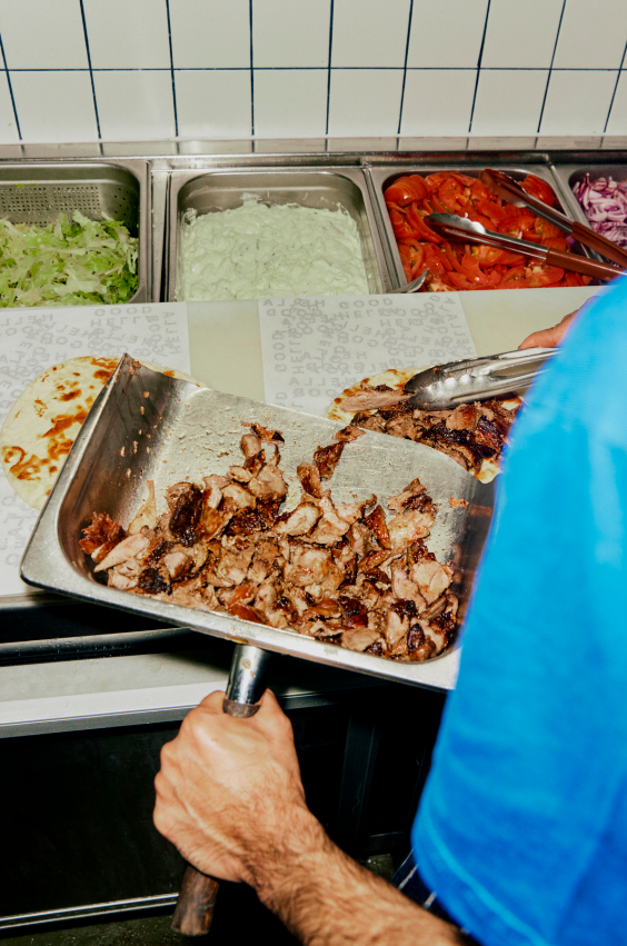 Person serving cooked pork from a metal tray onto a plate in a restaurant or food service setting, with salad ingredients like lettuce, lettuce with sauce, sliced tomatoes, and red onions in the background.