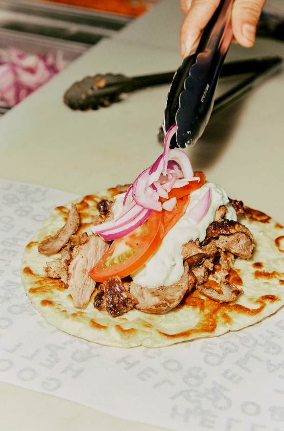 Person using tongs to place sliced onions on a taco with grilled meat, tomato, and sour cream on a soft tortilla.