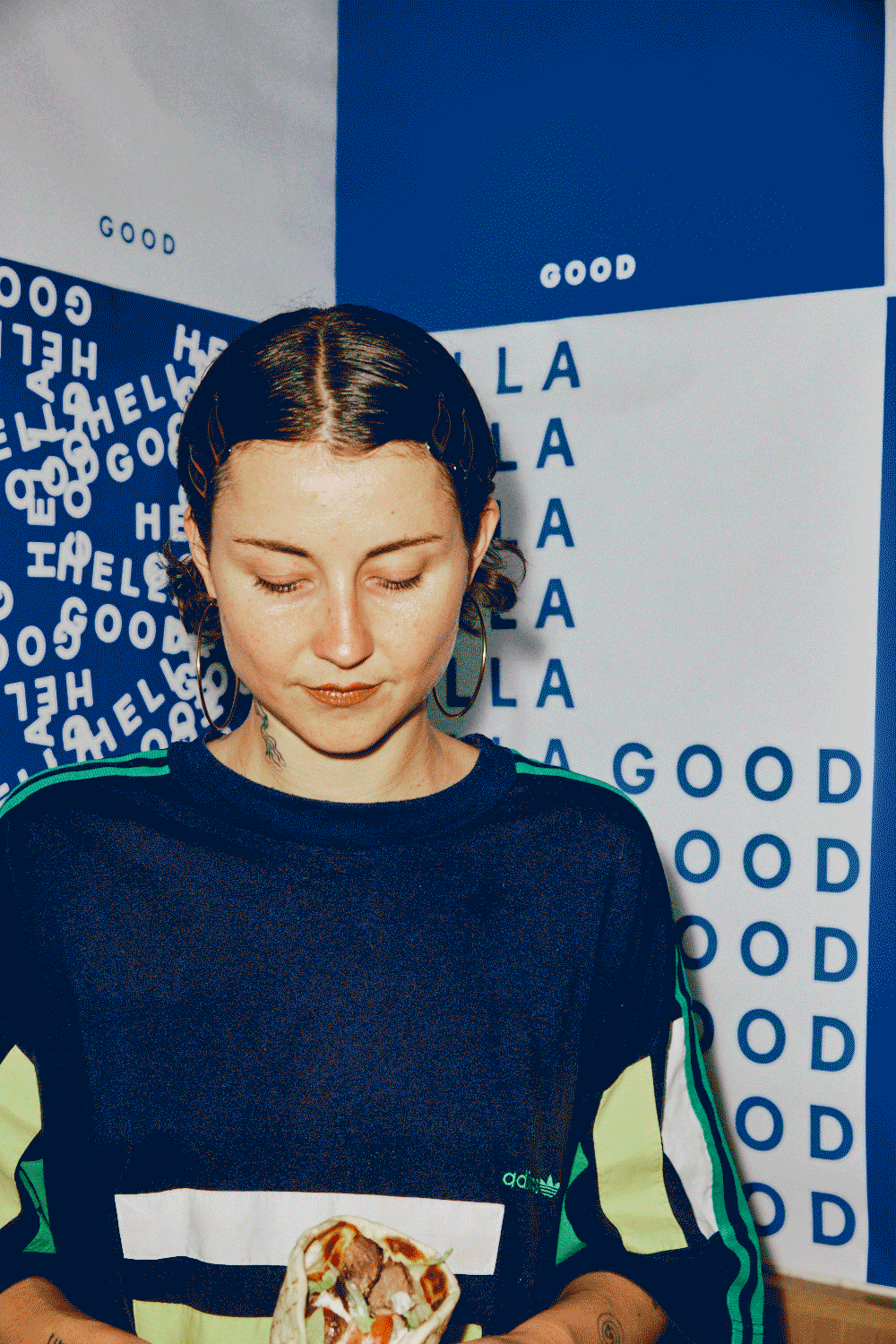 A young woman with dark hair and hoop earrings holding a slice of pizza with toppings. She is standing in front of a wall with blue and white text and patterns, including the words 'HELLO' and 'GOOD'.
