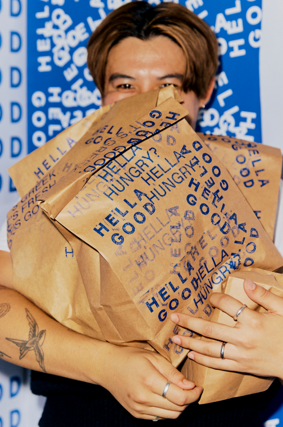 Person smiling and holding a large paper bag with blue text on it, in front of a blue backdrop with white text.