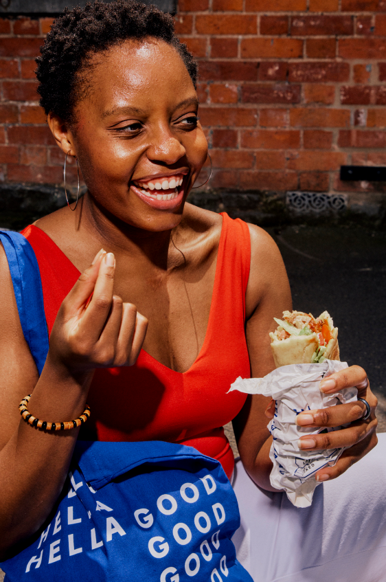 Woman with short curly hair wearing an orange tank top and hoop earrings, smiling and holding a sandwich wrapped in paper outdoors against a brick wall.