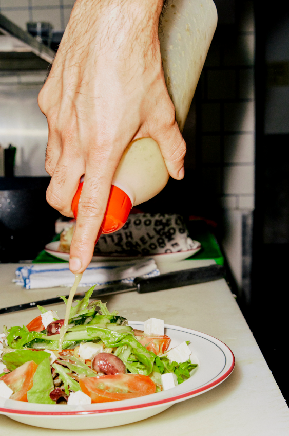 A hand drizzling creamy salad dressing from a squeeze bottle onto a mixed garden salad with greens, tomatoes, olives, and feta cheese on a white plate with red trim.
