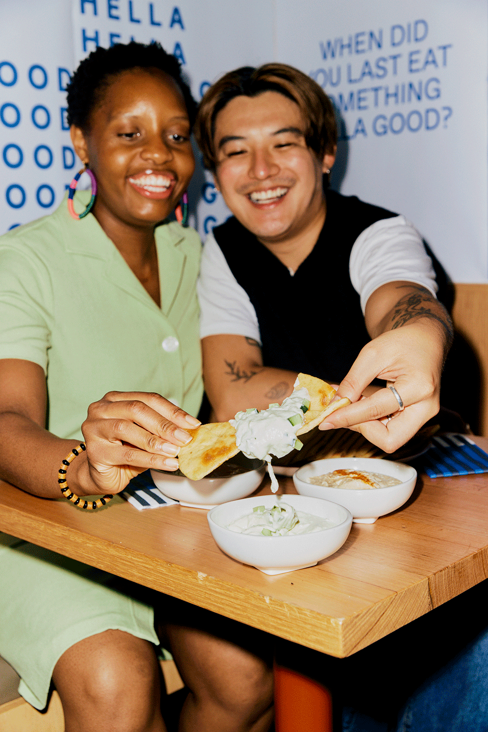 A woman and a man sitting at a restaurant table sharing a plate of food, with bowls of dips and a flatbread, smiling at the camera. The woman is wearing a green outfit, and the man is in a black and white shirt.