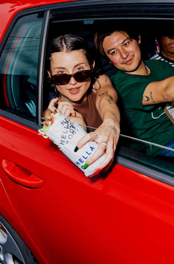 Two young women smiling from car windows, one wearing sunglasses and holding a burger wrapped in paper, with a red car door and window visible.