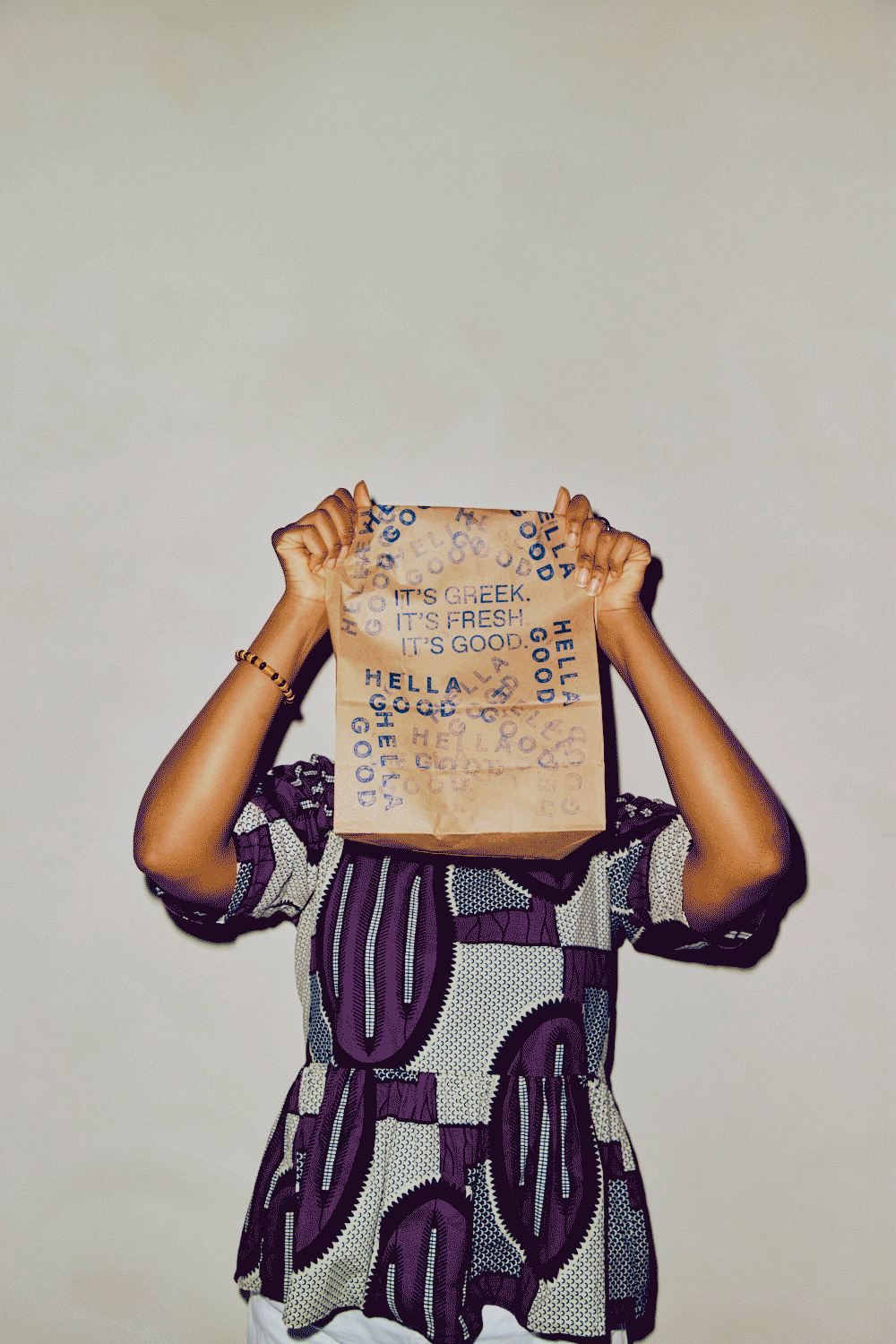 Person holding a paper bag with text covering their face, wearing a patterned purple, black, and white shirt, against a plain background.