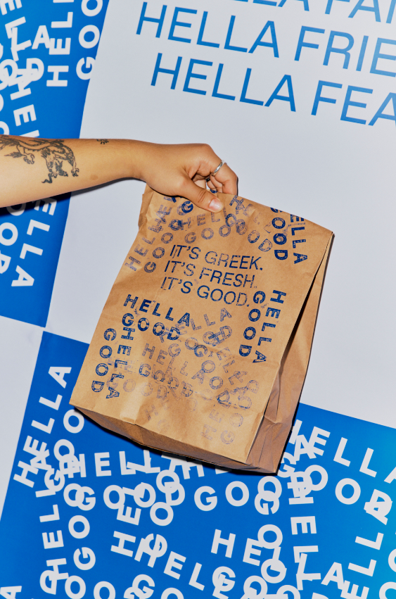 A hand holding a brown paper bag with blue printed text that says, "HELLO FRESH, IT'S GREEK, IT'S FRESH, IT'S GOOD," against a background of repeated blue and white text that says, "HELLO FRESH."