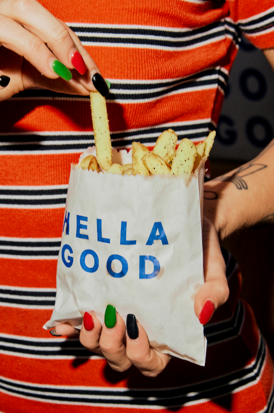 Person holding a white paper bag with French fries inside, one fry being held in hand. The bag has blue text saying "HELLO GOOD." Person has nails painted in red, green, and black. Person is wearing a red and black striped shirt.