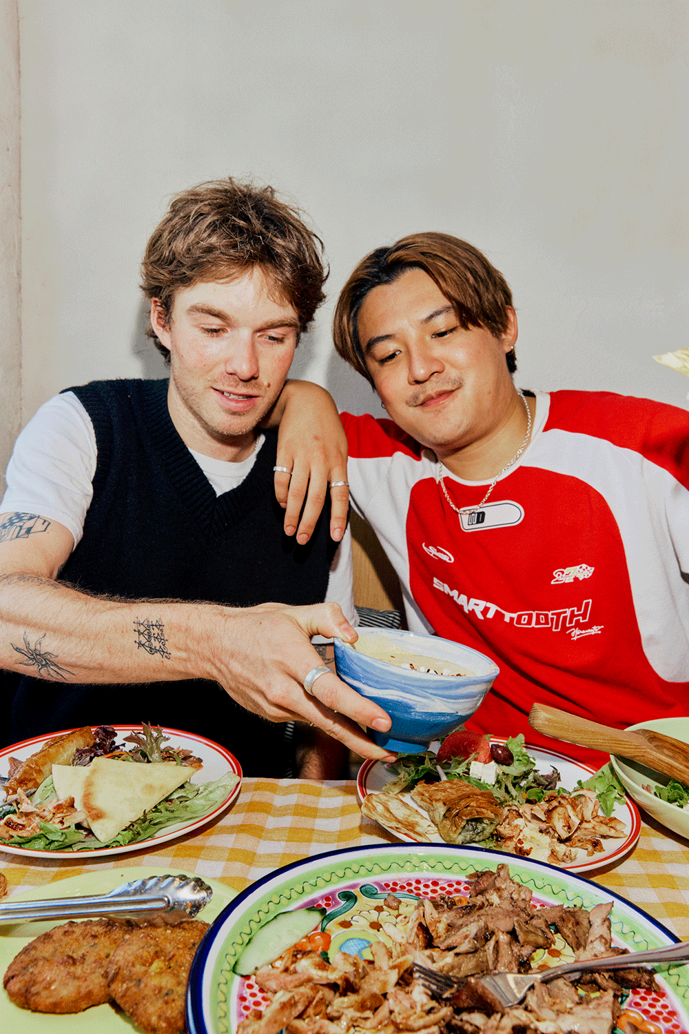Two men enjoying a meal together, with various dishes on the table including salads, grilled meats, and bread, sharing a bowl of food.