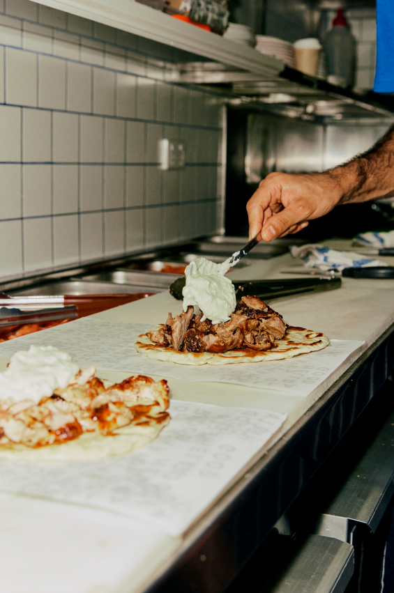 A chef adding whipped cream or a similar topping to a taco with meat and vegetables on a parchment paper-lined surface in a restaurant kitchen.