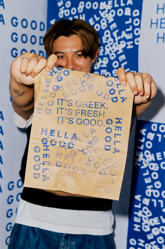 Person holding a paper bag with blue text that reads 'It's Greek. It's Fresh. It's Good', smiling behind it, with a background featuring matching blue and white text.