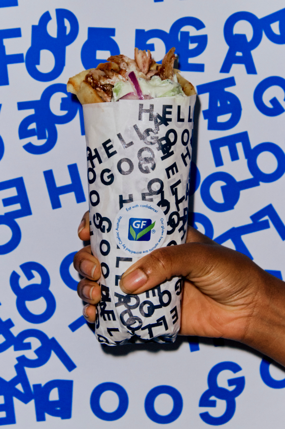 Hand holding a taco wrapped in branded paper with blue and black lettering, filled with grilled chicken, lettuce, and toppings, against a background with blue and white lettering.