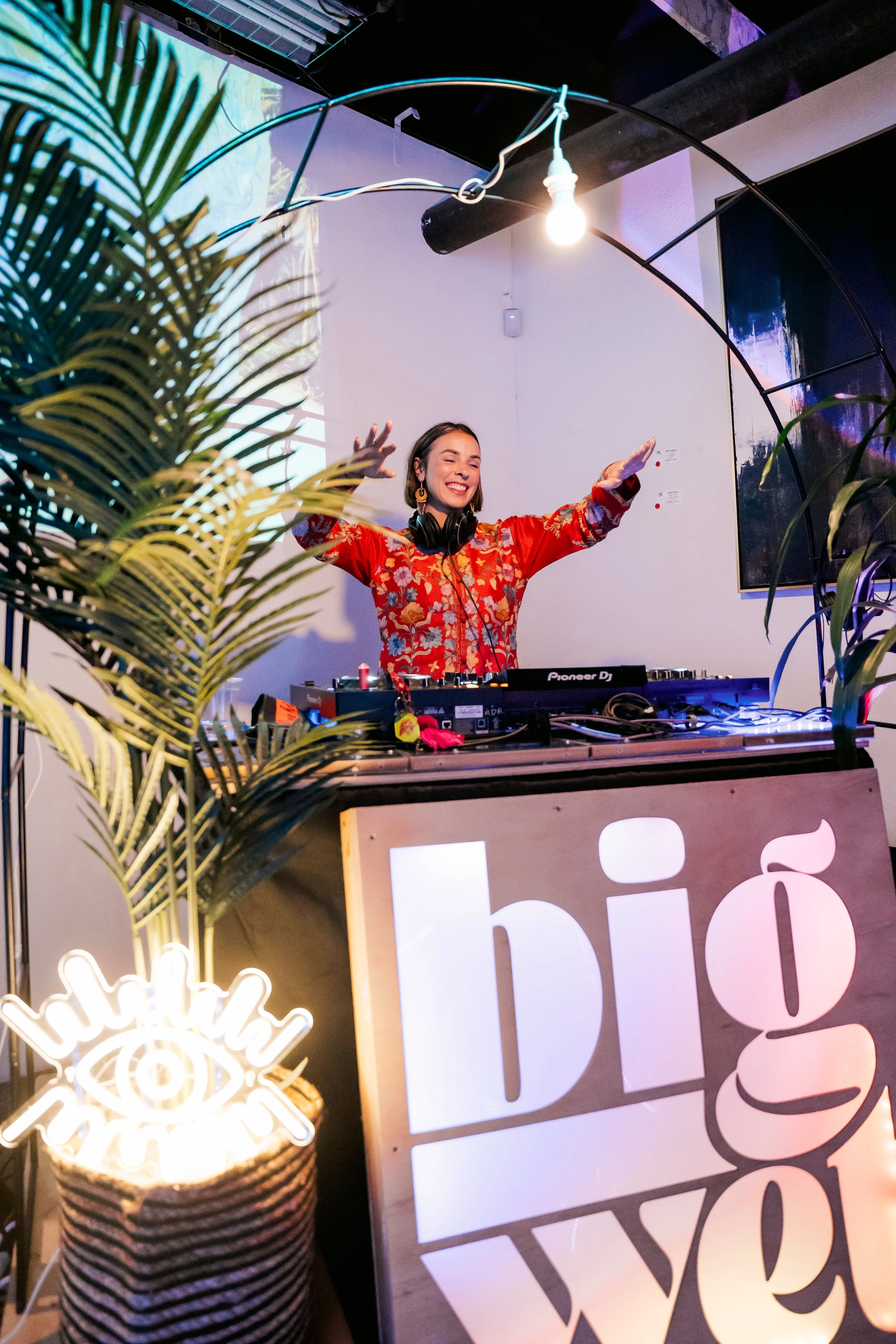 A female DJ wearing a red floral shirt with headphones around her neck, smiling and raising her hands while performing at a party. She is behind a DJ mixer, surrounded by tropical plants, with a neon sign reading "eye" and a wooden sign that says "bi