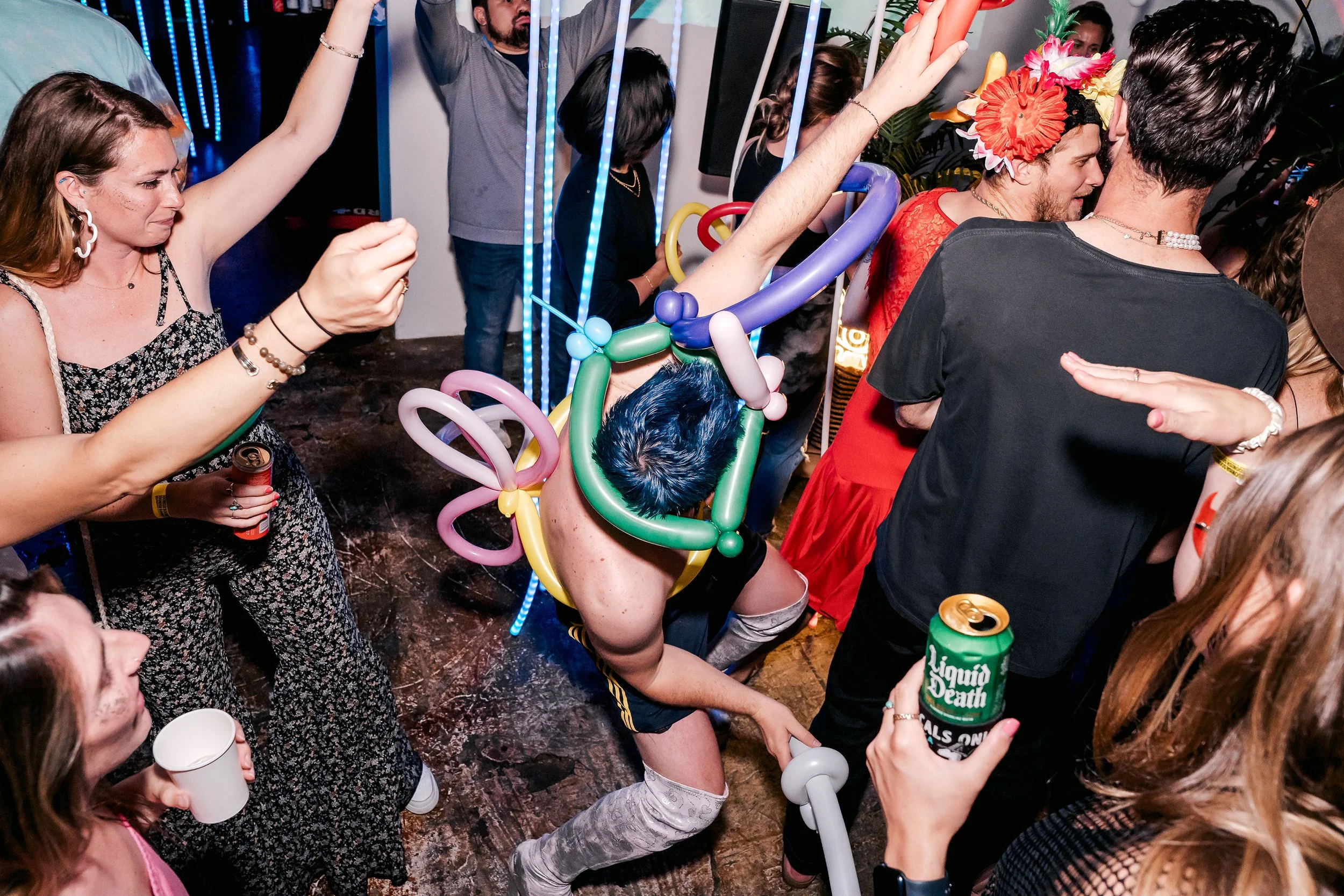 Party scene with people dancing, some holding drinks, one person wearing balloon sculptures and colorful flower headgear, others in casual attire, in a lively indoor setting.