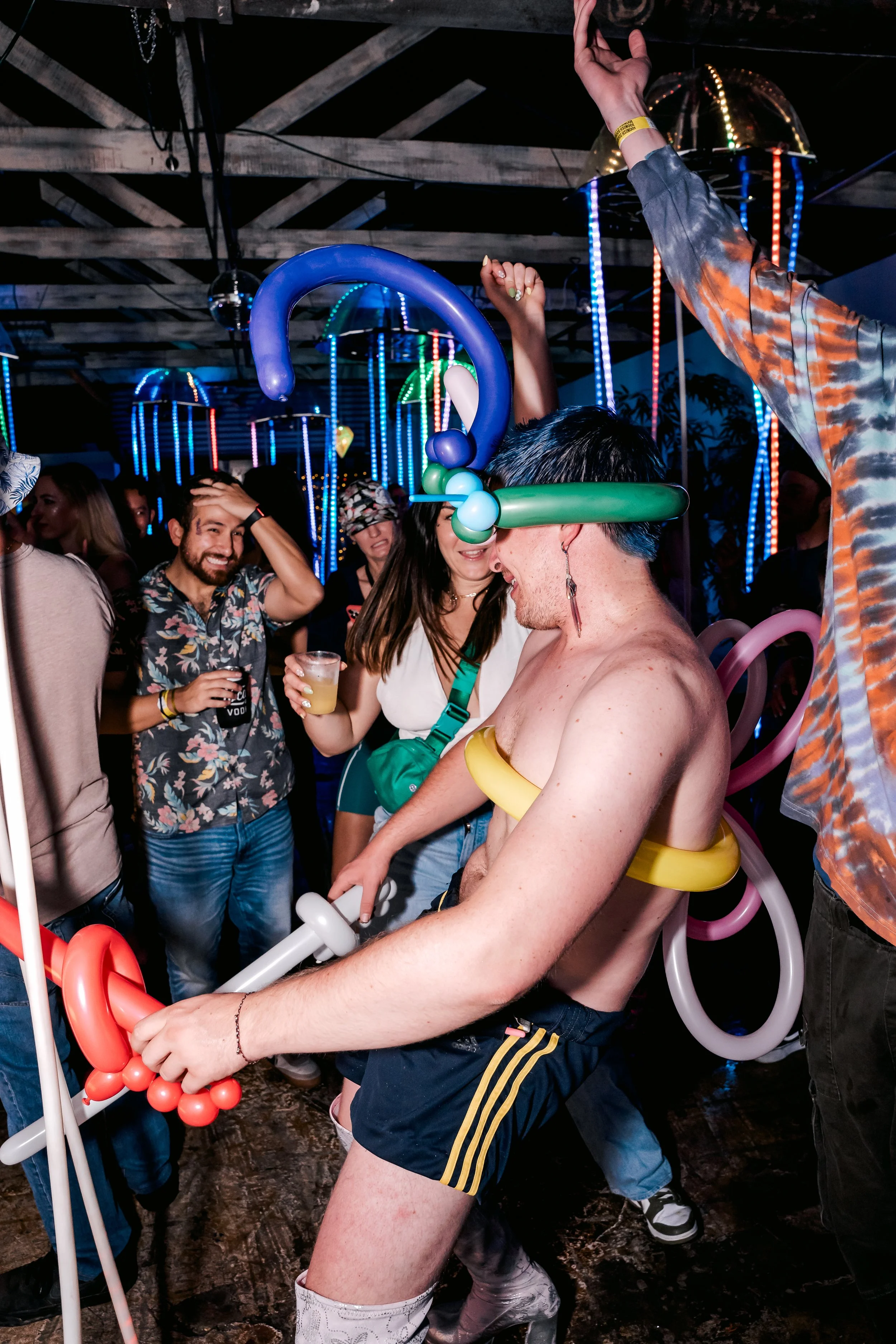 People celebrating at a party, wearing balloon hats and accessories, with colorful lights and a dark, rustic ceiling in the background.